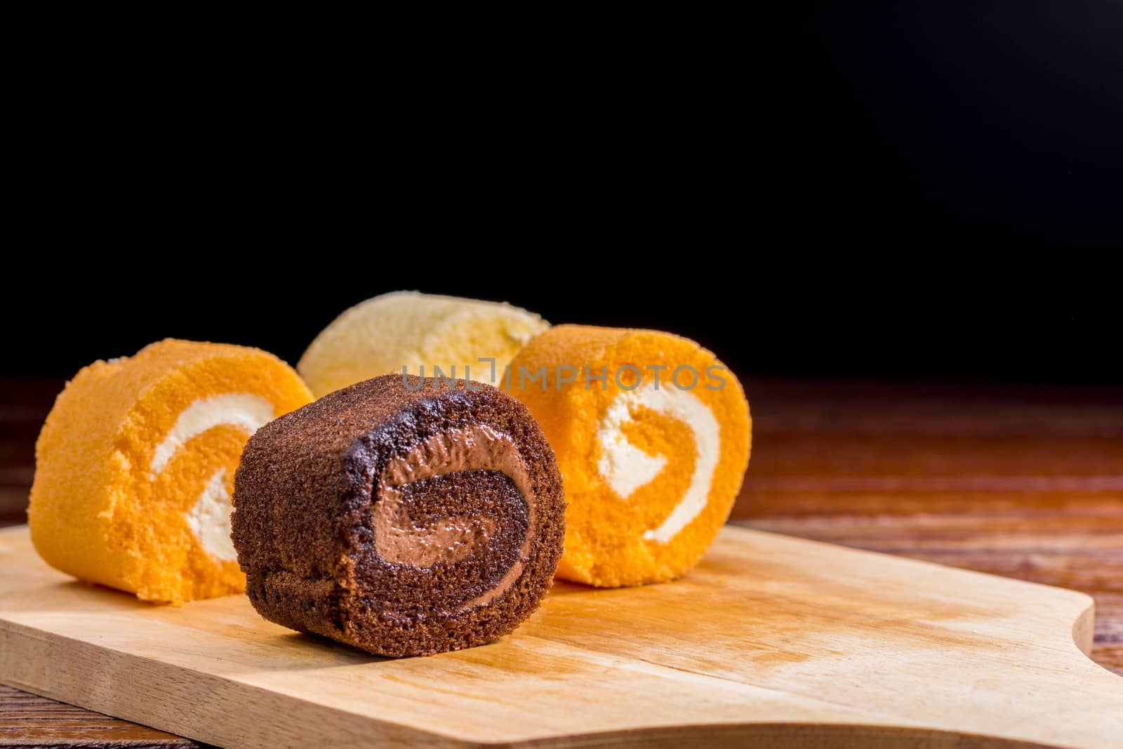 Close up delicious chocolate, vanilla and orange roll cake with whipping cream on  a wooden chopping board and table at the home kitchen with a black background and copy space. Homemade bakery concept.