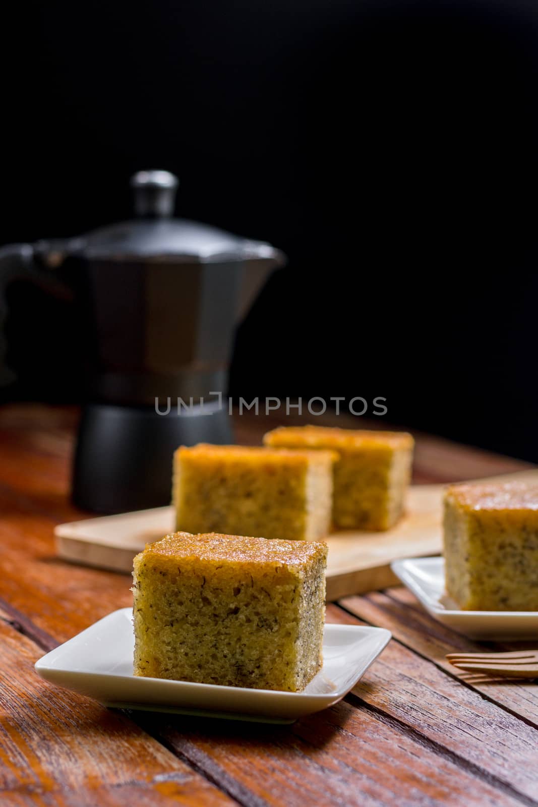 Close up square cut of homemade sweet and solf banana cake on wooden chopping board and fork on table with solf focus black moka pot. Delicious and healthy bakery.