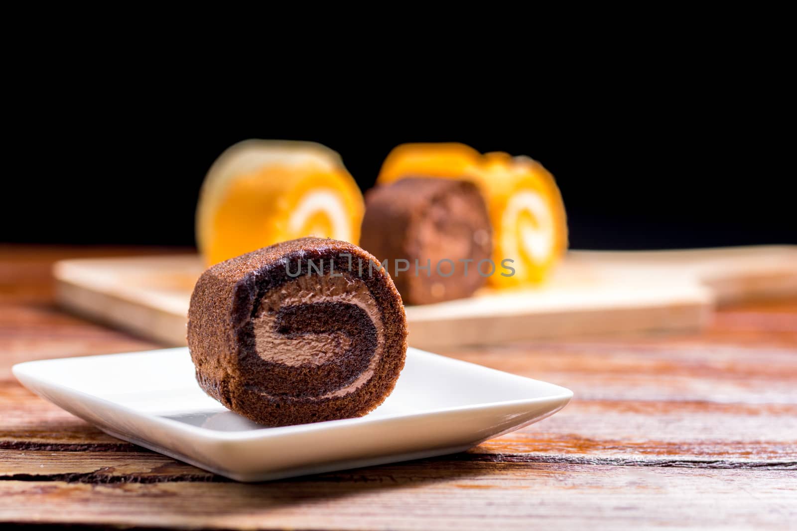 Close up delicious chocolate roll cake with whipping cream in a square white plate on a wooden table at the home kitchen with a black background and copy space. Homemade bakery concept.