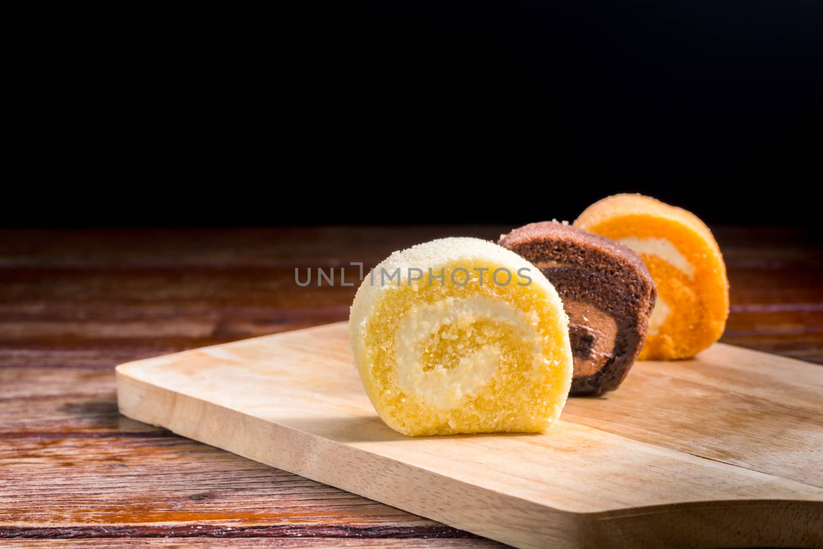 Close up delicious chocolate, vanilla and orange roll cake with whipping cream on  a wooden chopping board and table at the home kitchen with a black background and copy space. Homemade bakery concept.