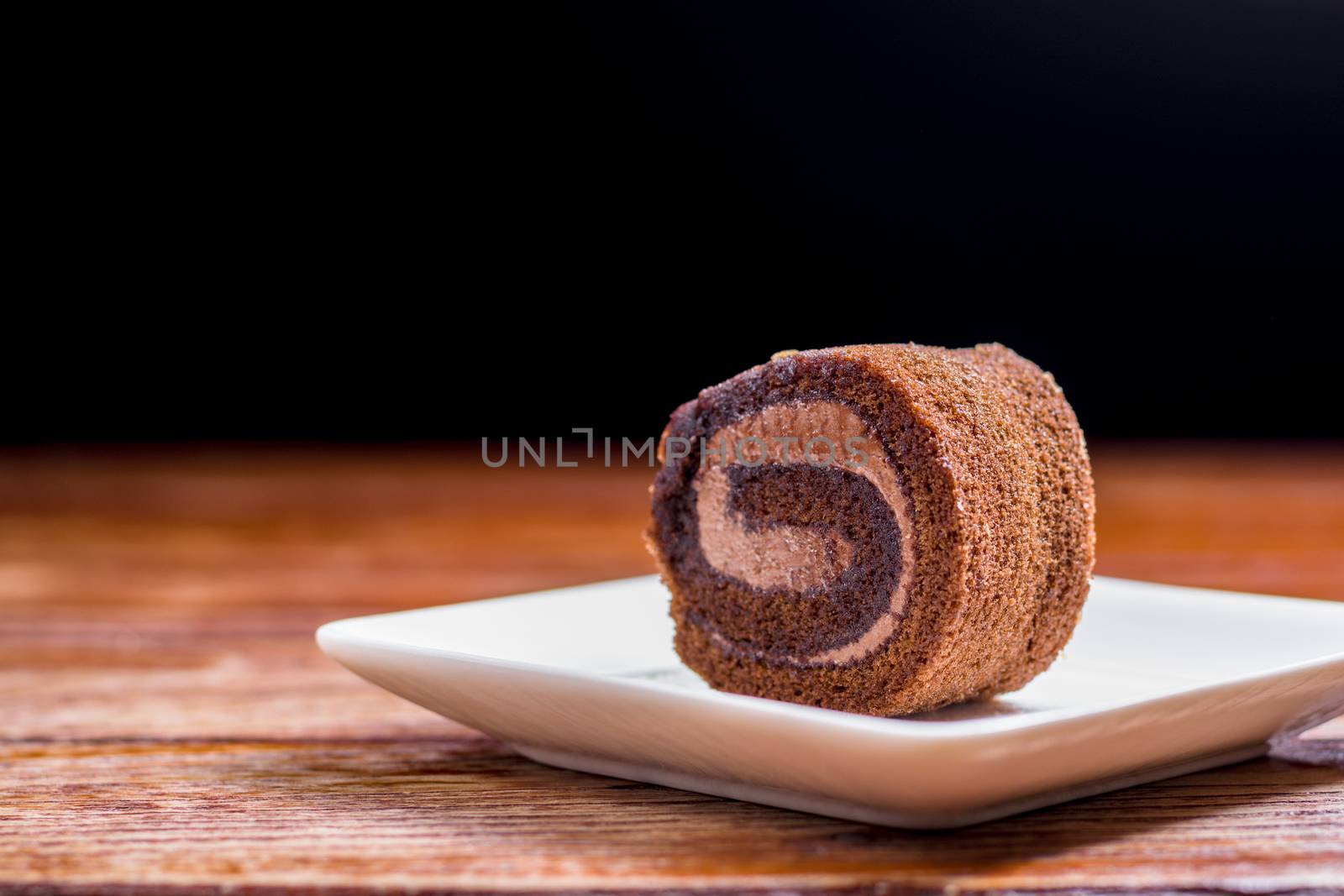Close up delicious chocolate roll cake with whipping cream in a square white plate on a wooden table at the home kitchen with a black background and copy space. Homemade bakery concept.