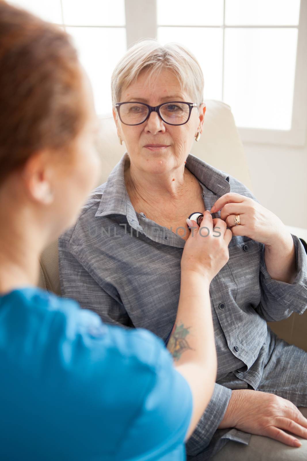 Back view of female nurse using stethoscope in nursing home to listen old woman heart beat.