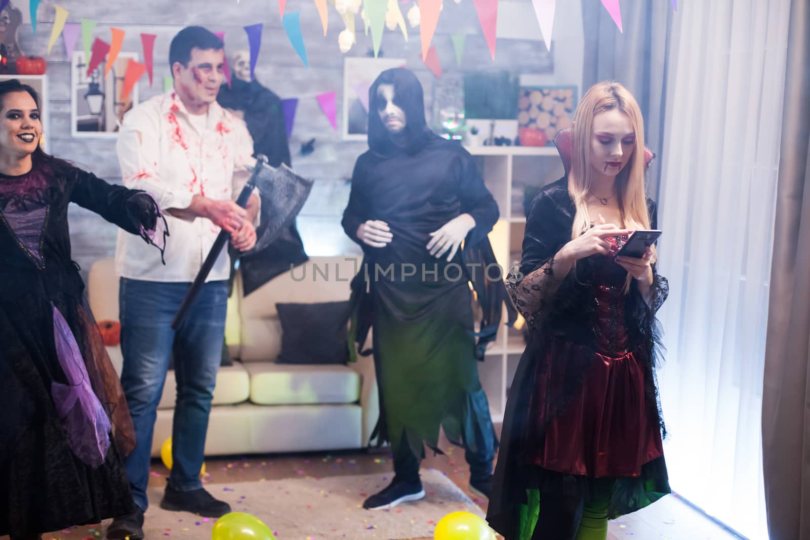 Beautiful young woman dressed up like a vampire with bloody lips using smartphone at halloween party.