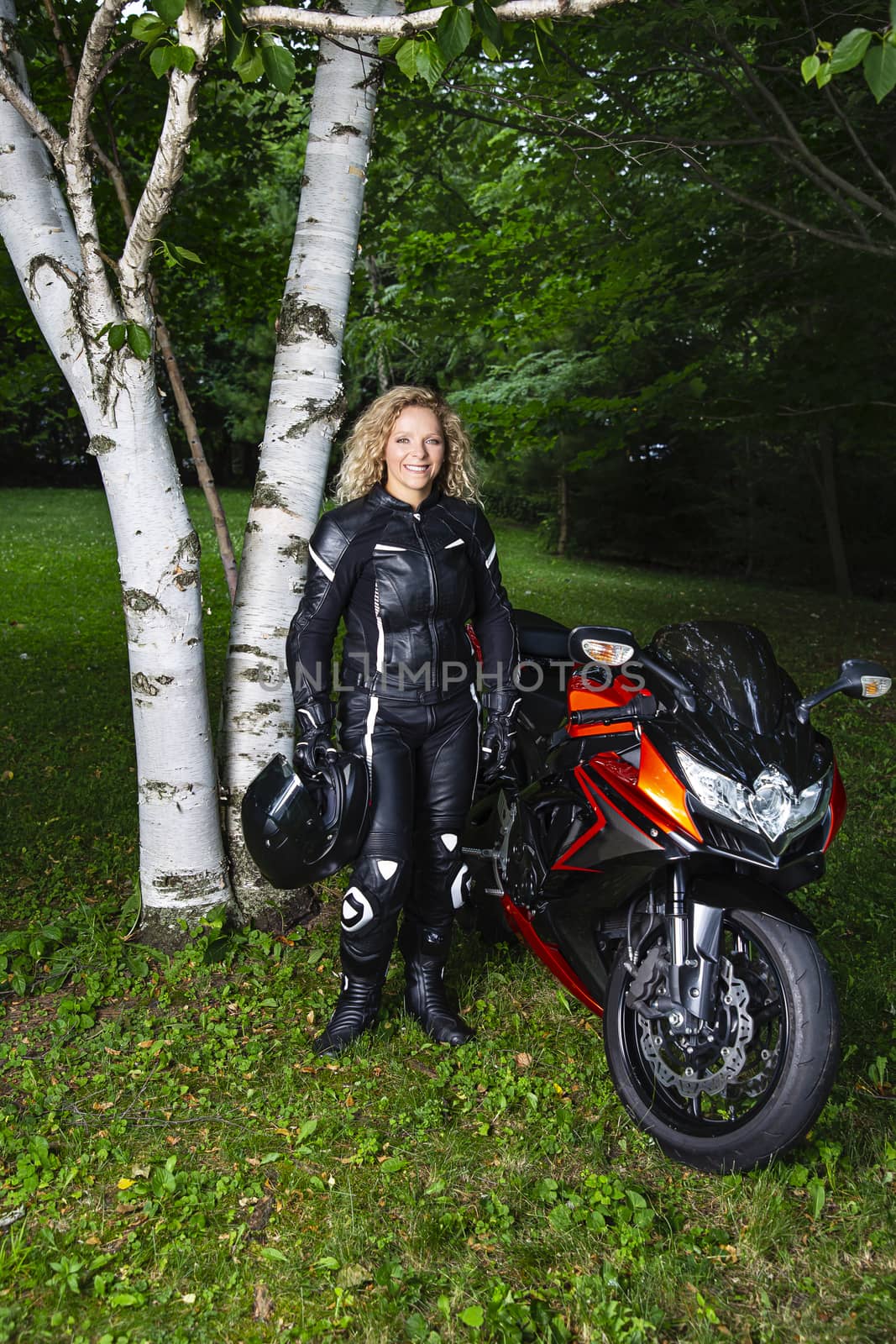 twenty something blond woman, wearing full motocycle gear, standing beside sport motocyle, in a park