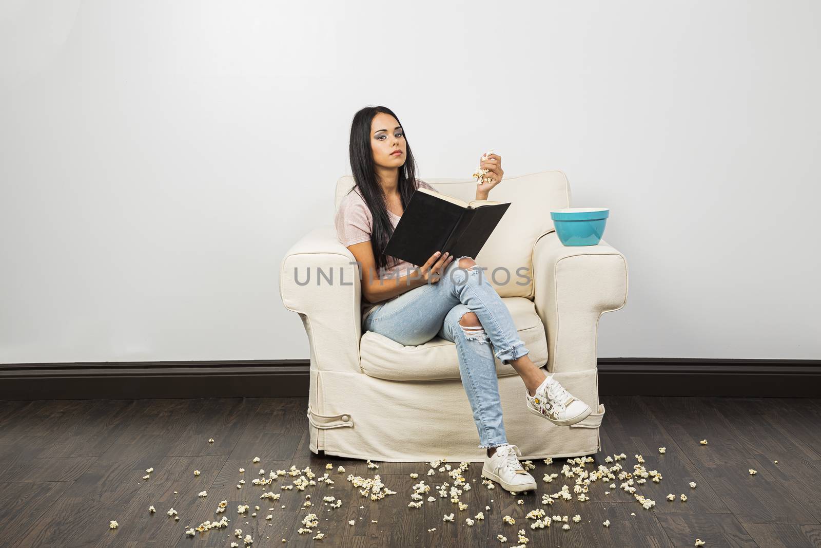 young woman, making a mess of popcorn around a white couch, while reading a hard cover book