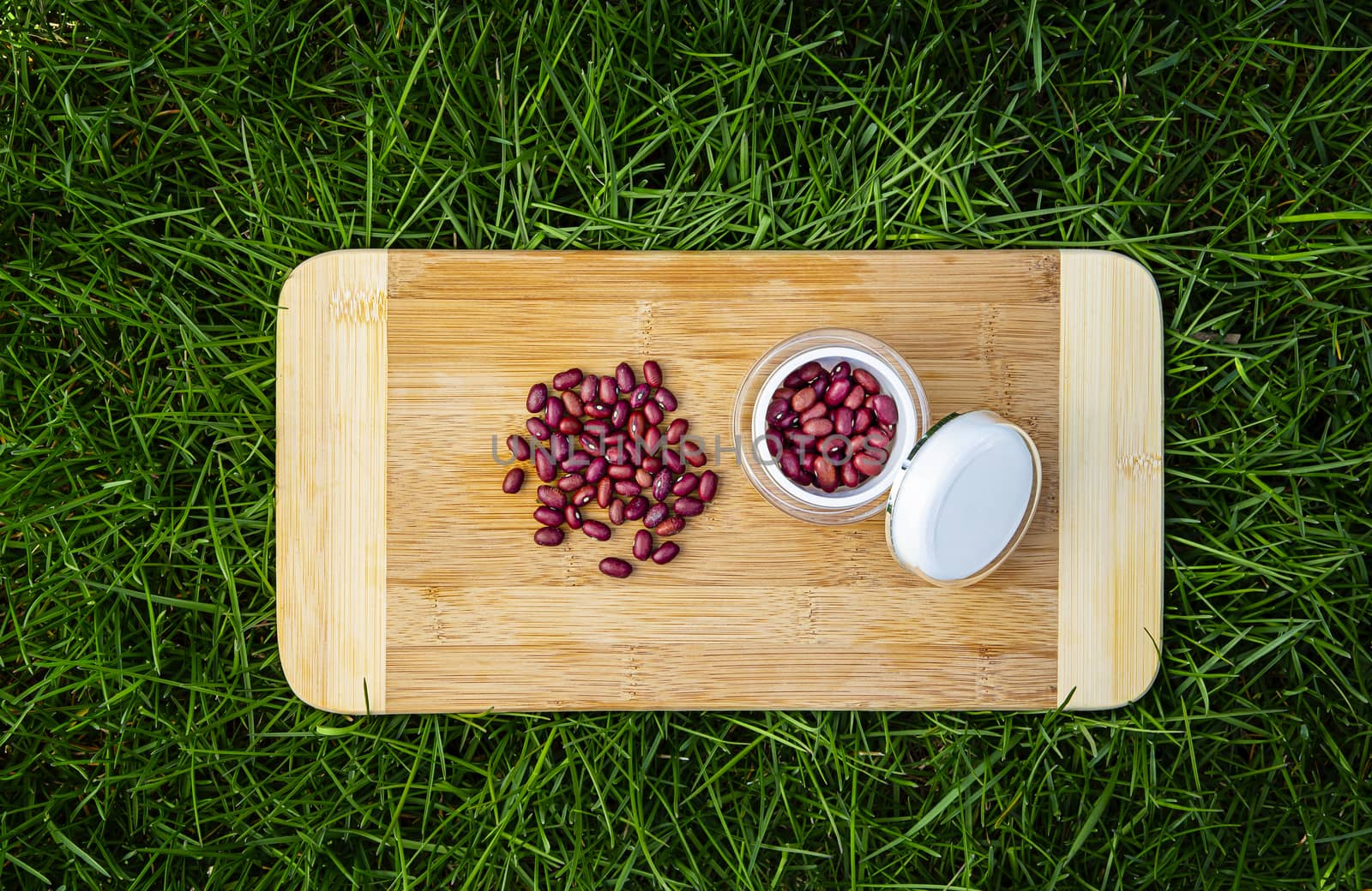 pile of red kidney beans on a wood cutting board, in green grass and small beauty cream container filled with red kidney bean
