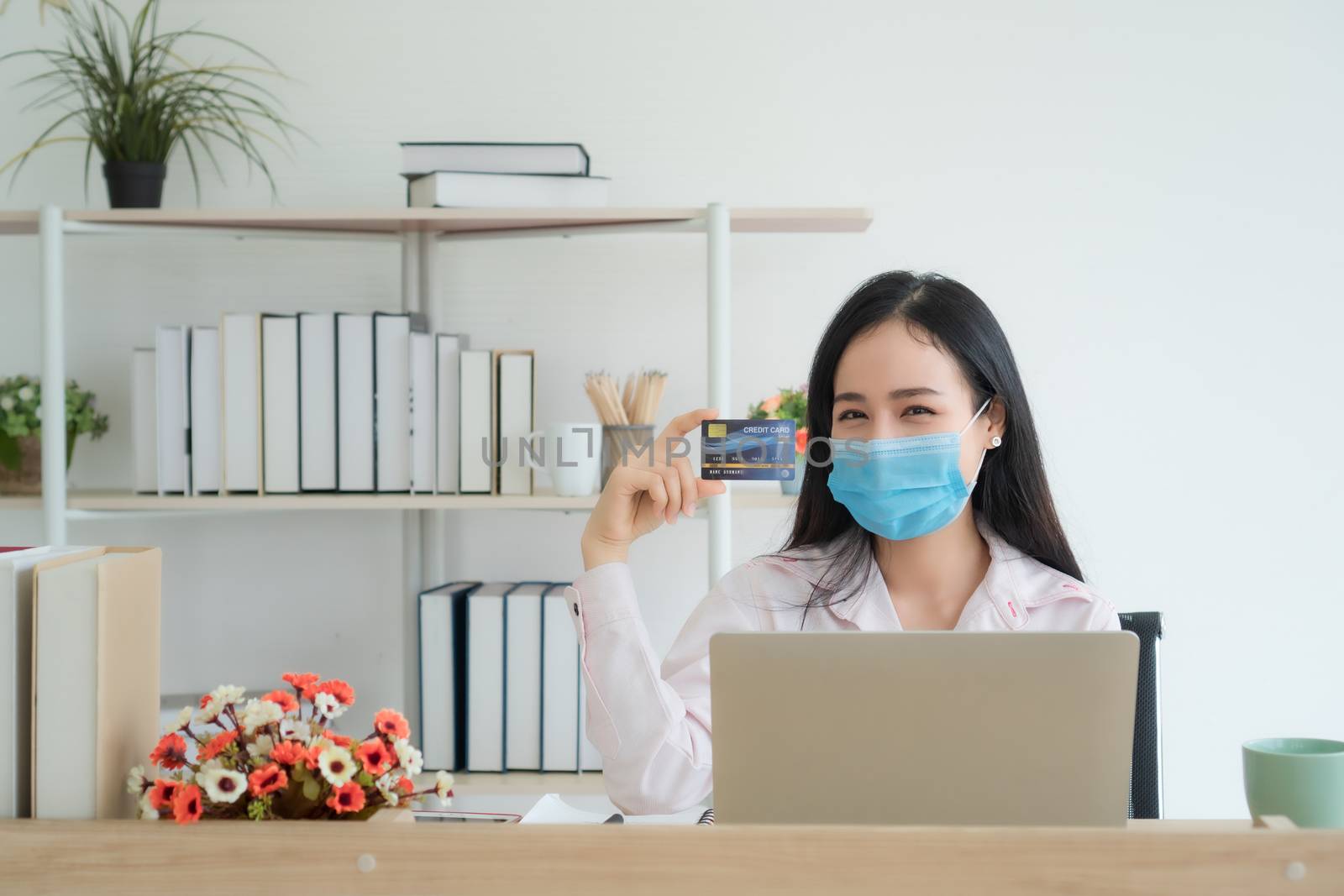A beautiful Asian woman is working at home happily. She is using a laptop to work. She wore a mask to prevent the outbreak of the virus. She held a credit card in her hand.( Mock up  credit card )
