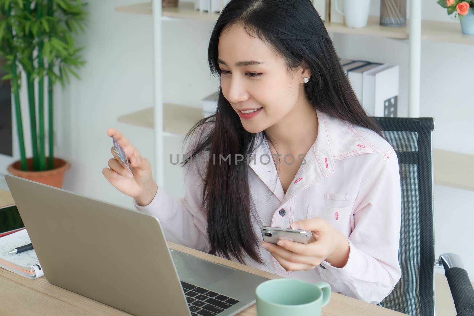 Work from home - Concepts. A beautiful Asian woman is working at home happily. She is using a laptop to work. She holds a credit card. And the smartphone is in the hand.( Mock up  credit card )