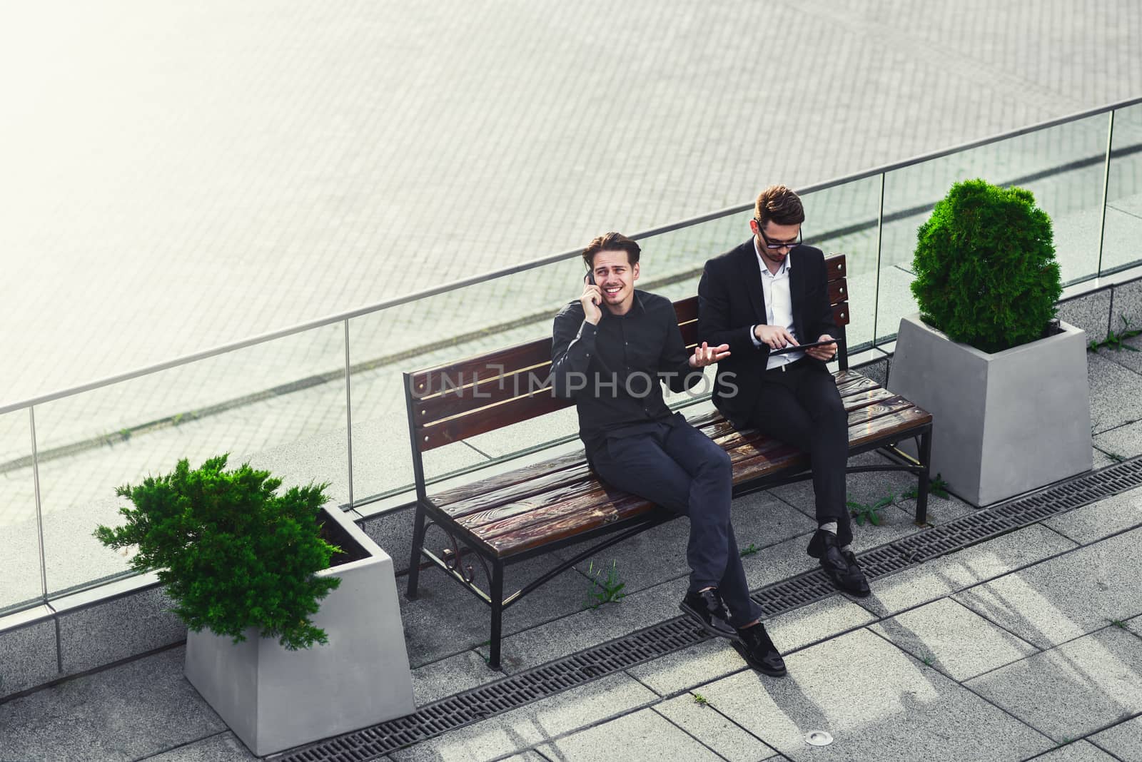 Two Businessman's have a conversation on street bench busy by phone and tablet.