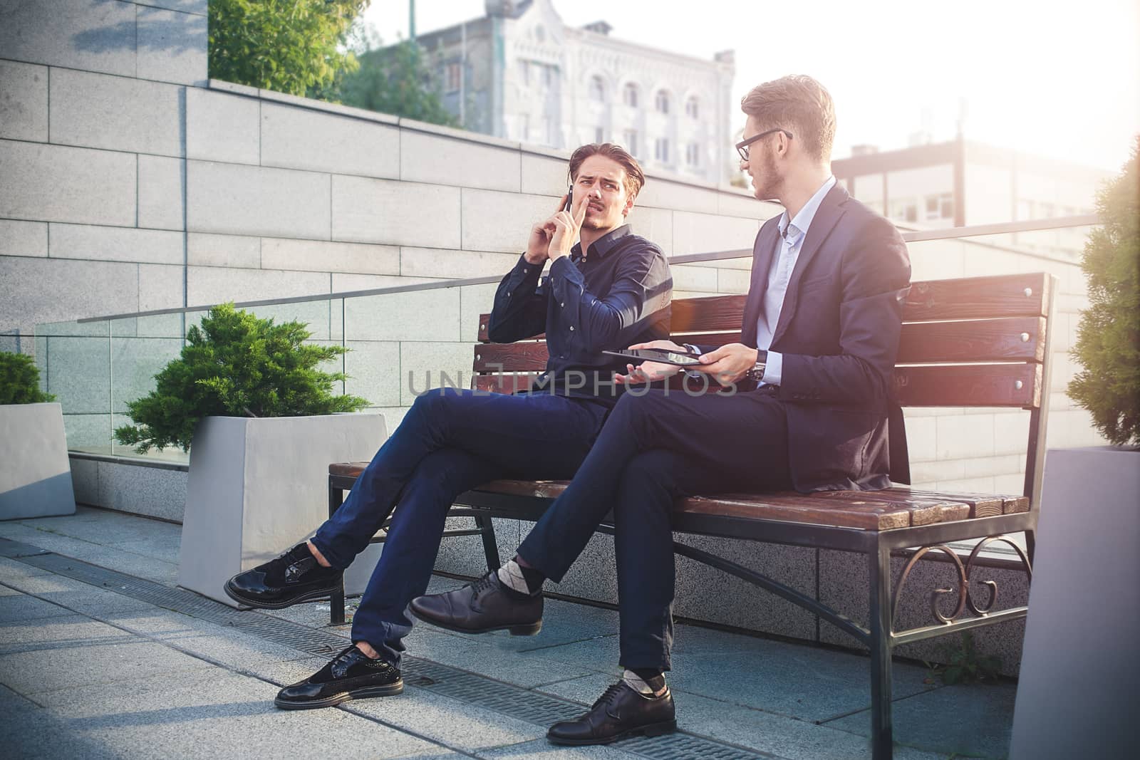 Two Businessman's have a conversation on street bench busy by phone and tablet.