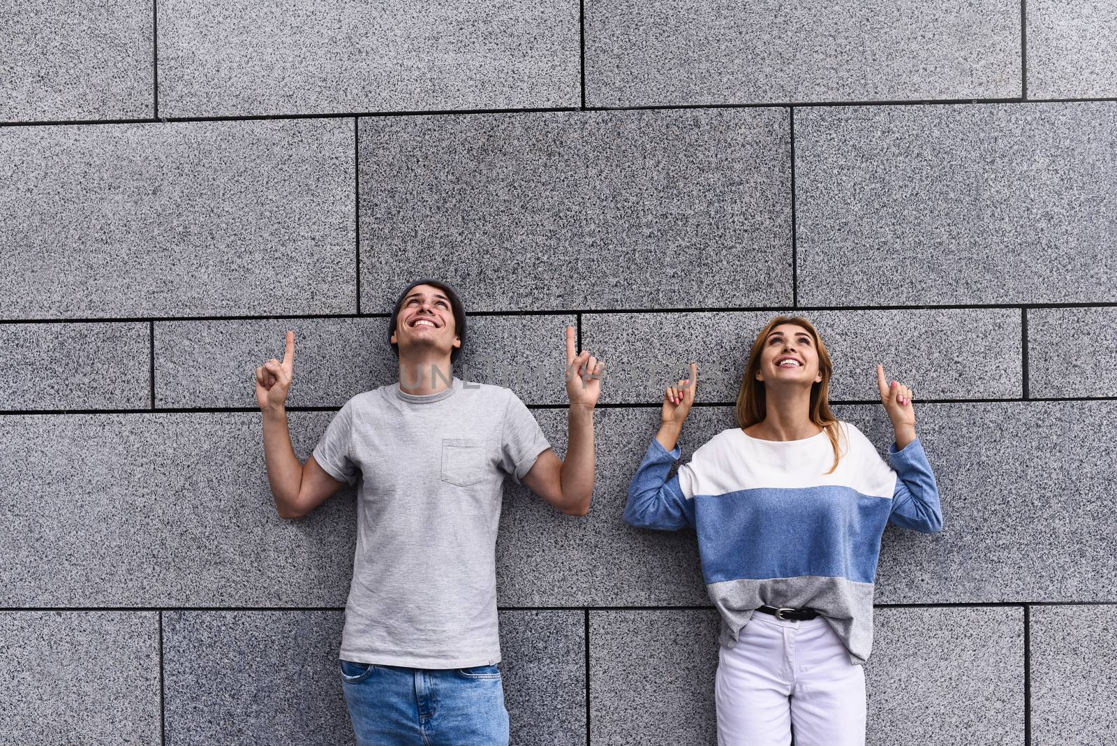 Cheerful couple at the street, show a big sale, or nice offer. Advertise concept. People, travel and tourism - on city street and couple show with their fingers something over gray wall background.