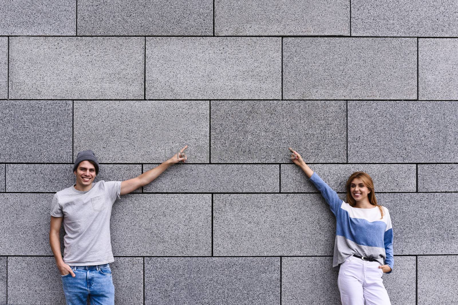 Cheerful couple at the street, show a big sale, or nice offer. Advertise concept. People, travel and tourism - on city street and couple show with their fingers something over gray wall background.