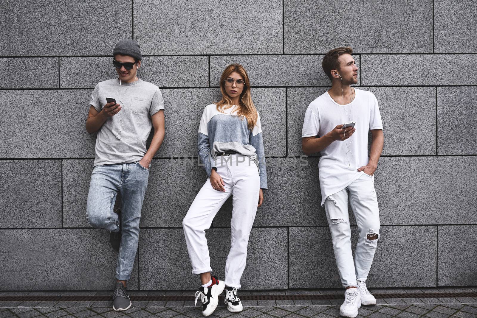 Fashion portrait of Three best friends posing at street, wearing stylish outfit and jeans against gray wall . by Nickstock