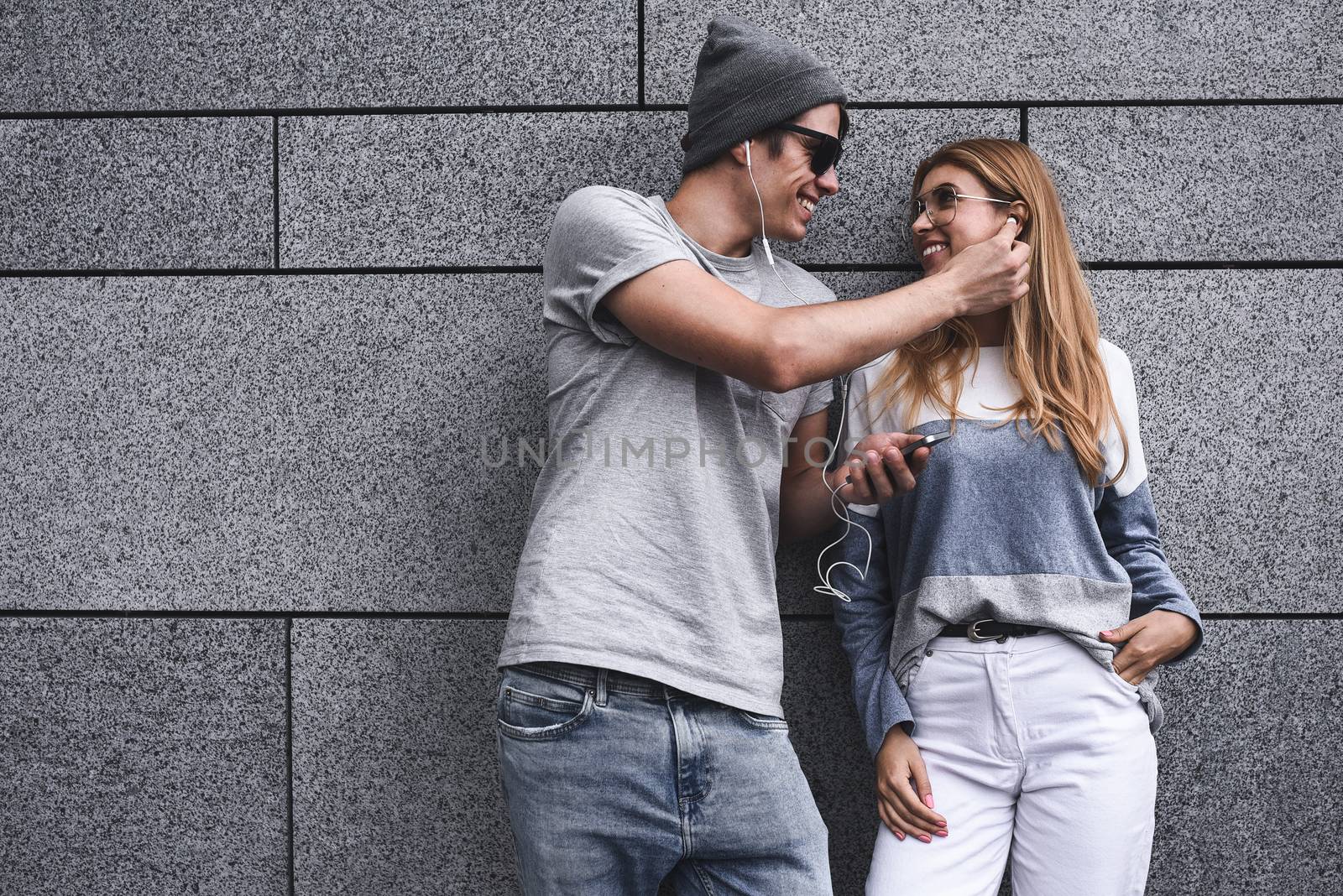 Young attractive couple listening to music on the same pair of headphones, dressed in stylish clothes against a background of a gray wall.