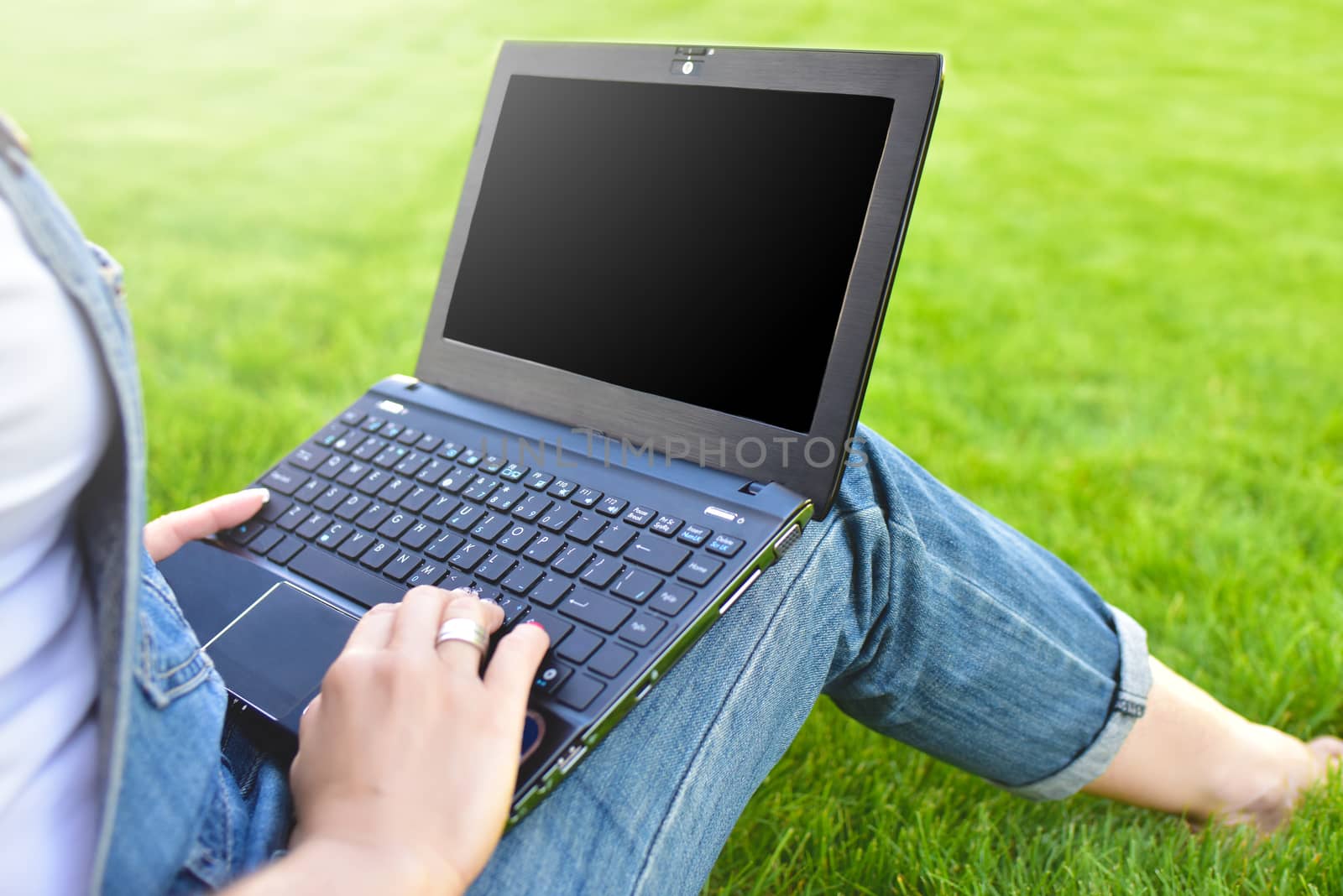 Close view of woman sitting on green grass in park with laptop on legs, spending summer day working outdoor, using laptop and wireless Internet for online work.