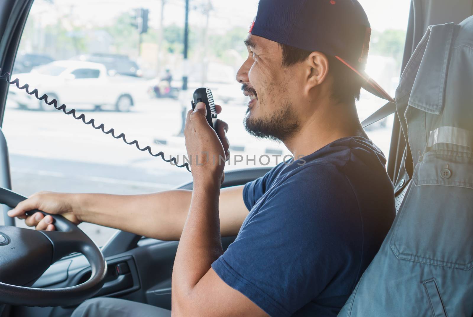 An Asian male truck driver is driving down the road with caution. He is using radio to communicate with the call center.