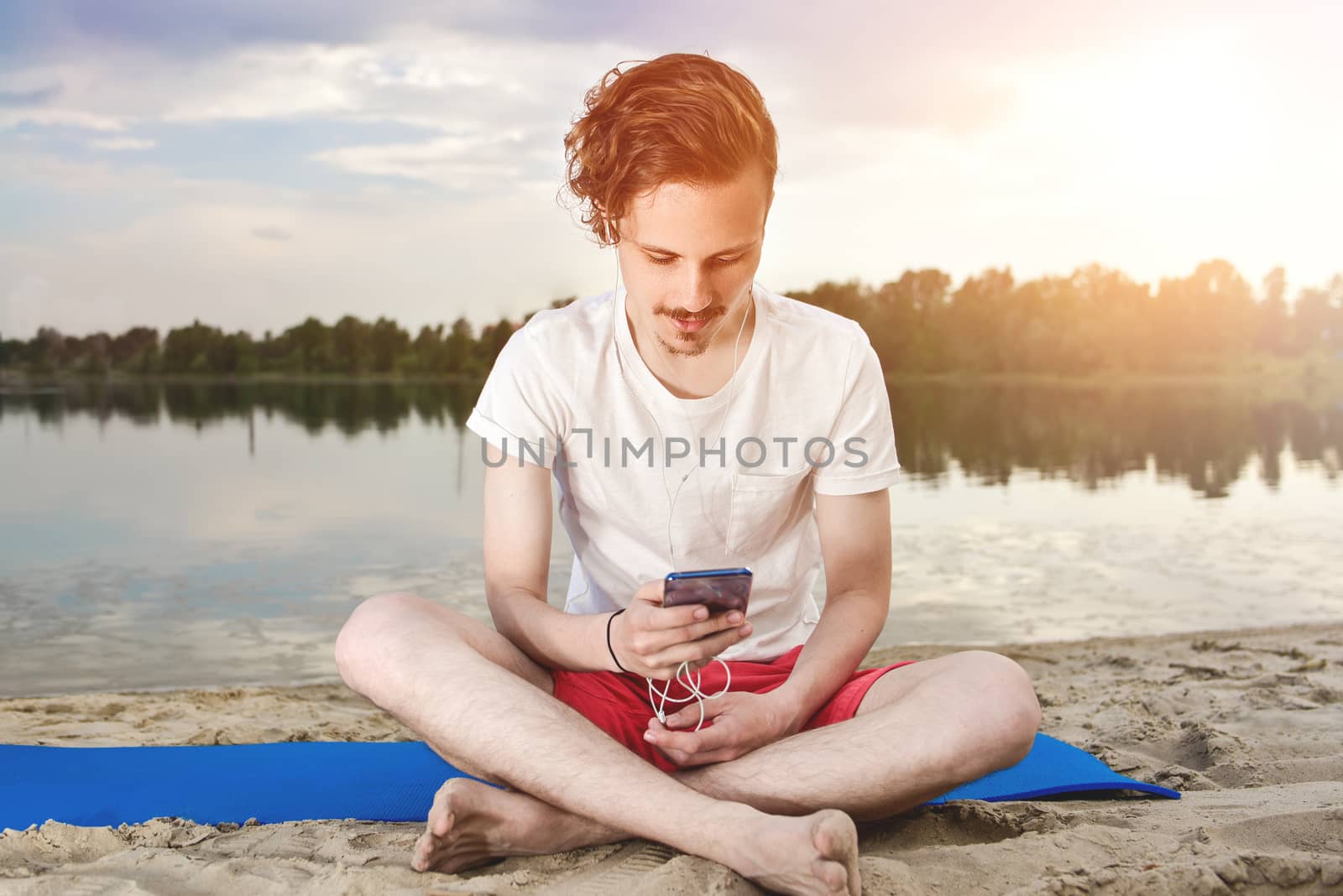 Man is sitting on the beach at lake and writing a sms on phone. good mood. beautiful view