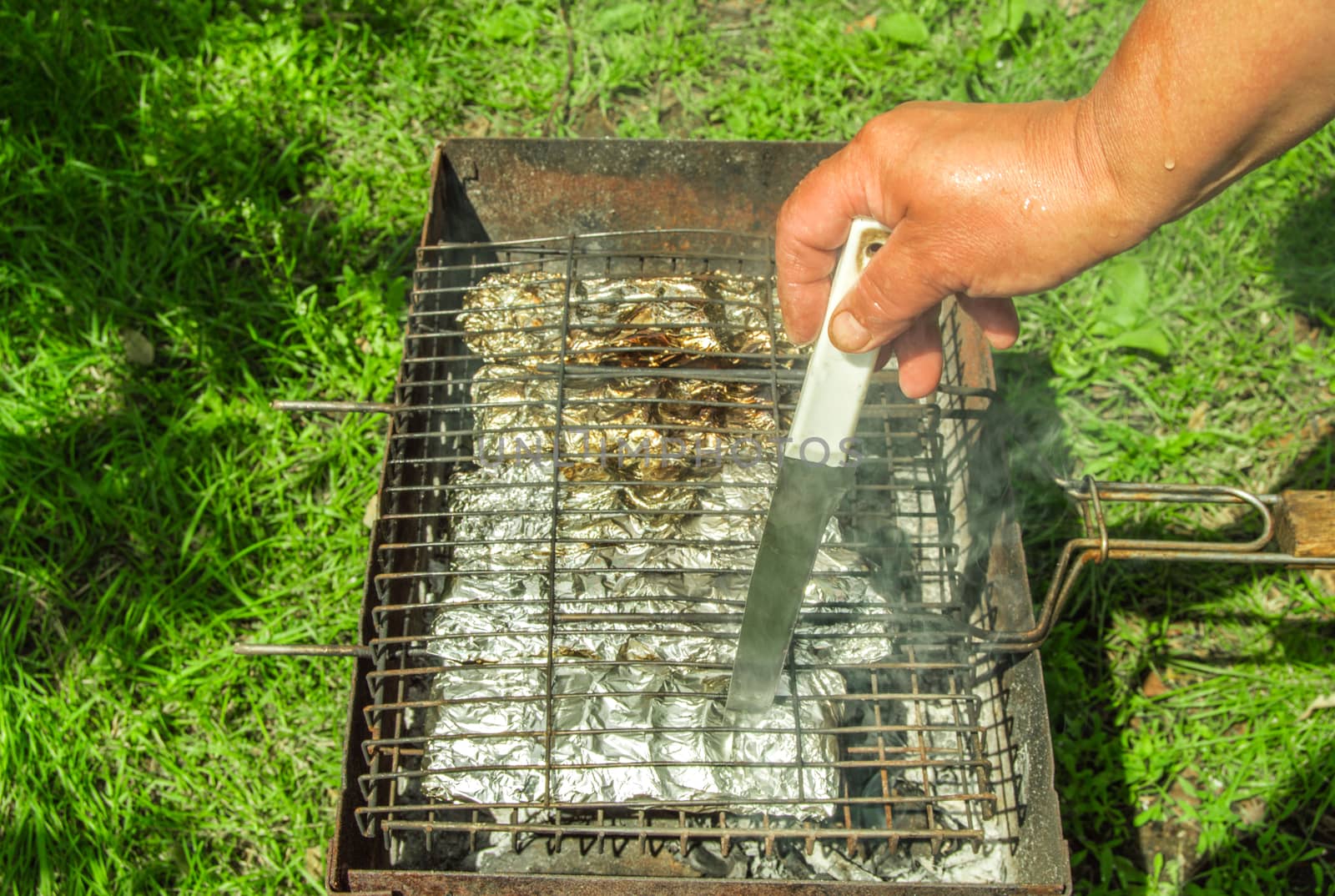 The hand of a person checks the readiness of food with a knife. Cooking baked vegetables in foil on the grill. The concept of healthy eating, outdoor.