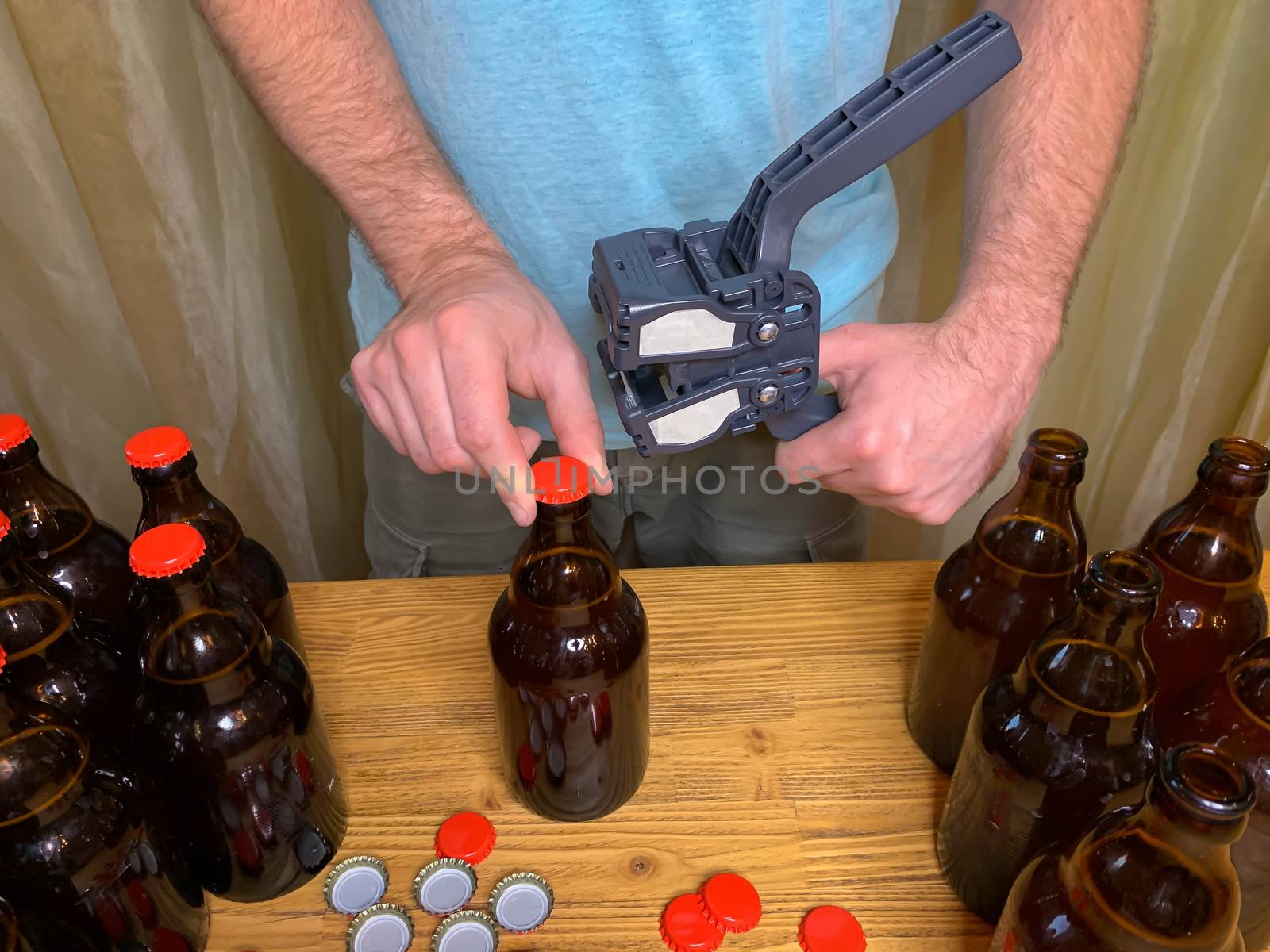 Craft beer brewing at home, man closes brown glass beer bottles with plastic capper on wooden table with red crown caps. Horizontal stock image