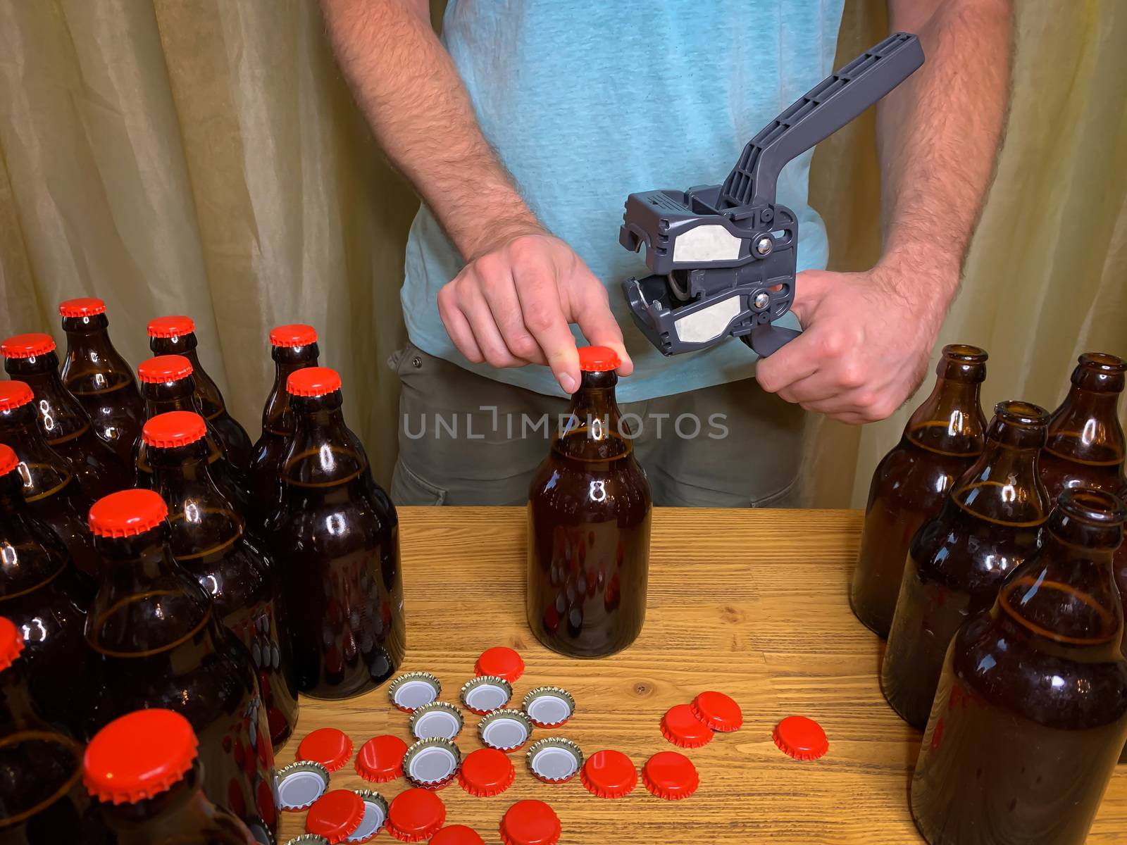 Craft beer brewing at home, man closes brown glass beer bottles with plastic capper on wooden table with red crown caps. Horizontal stock image