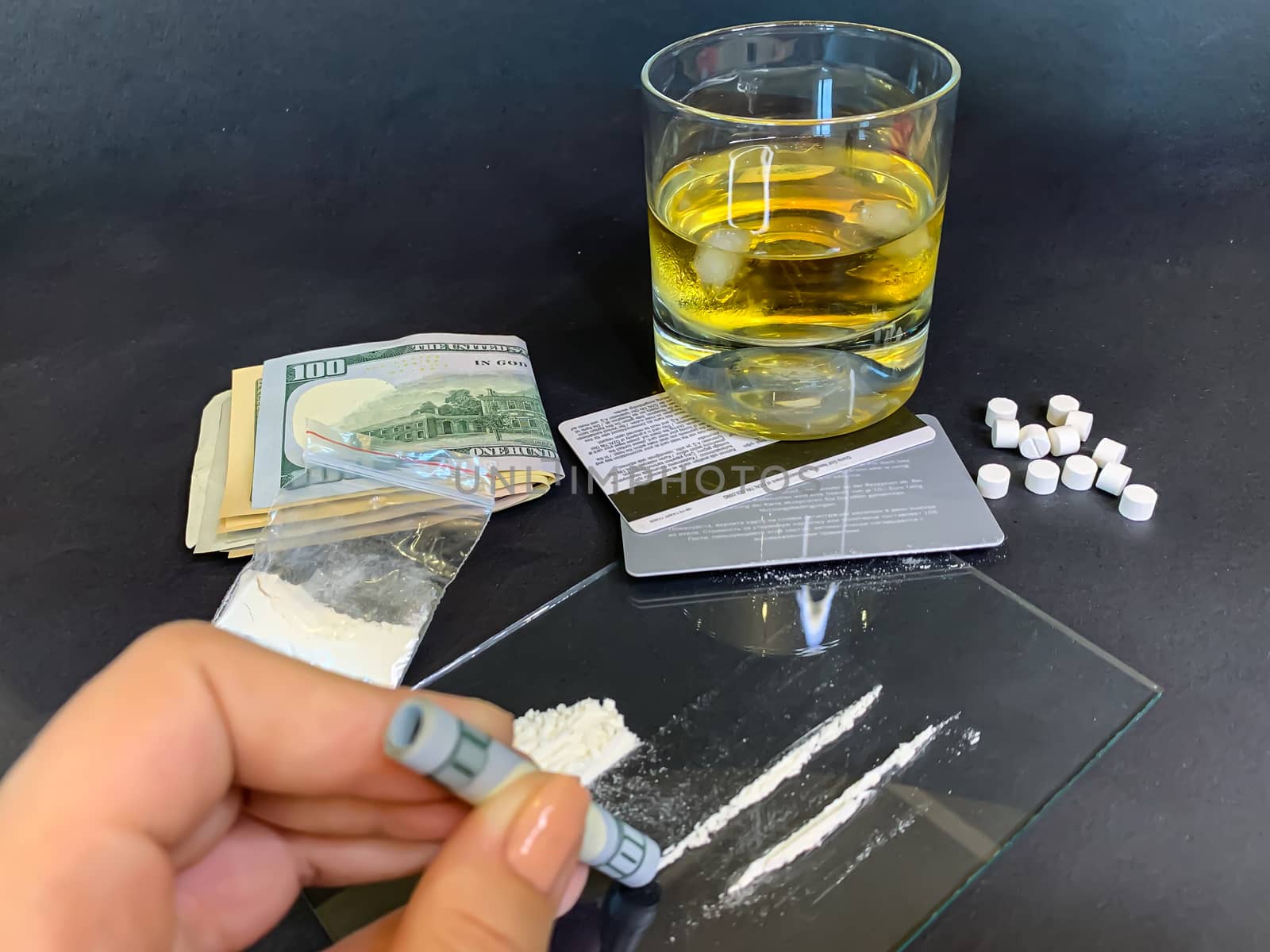 A female's hand holding rolled 100 us dollars banknote near two lines of cocaine. Plastic bag with illegal drugs, tablets, money and a glass of whiskey on background. Drug addiction. Stock image.