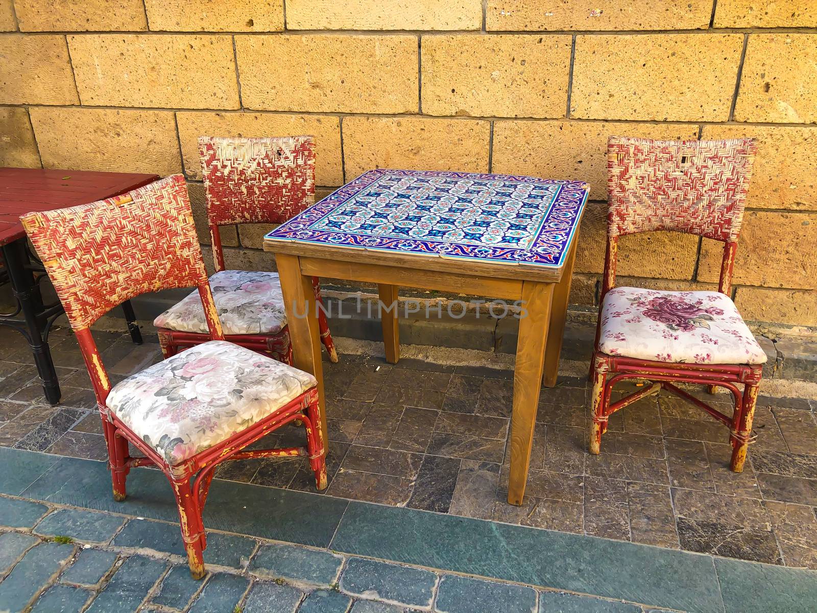 Antique table with blue mosaic tiles and three red old chairs near stone wall