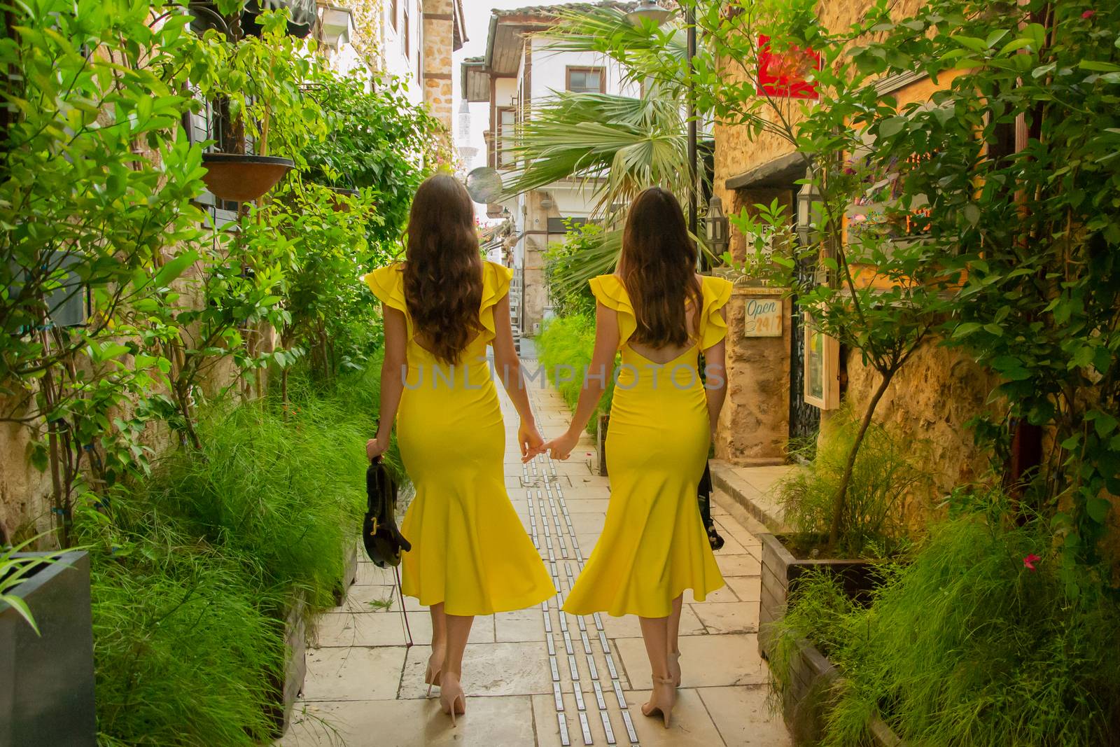 Two young girls with long dark hair are holding violins in their hands and walking down the historical street of old town of Antalya Kaleici, Turkey. Stock image, view from the backside. 