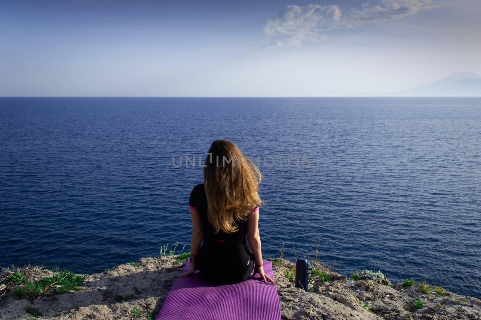 Beautiful young happy lonely girl sitting on rock and watching blue Mediterranean sea and relaxing