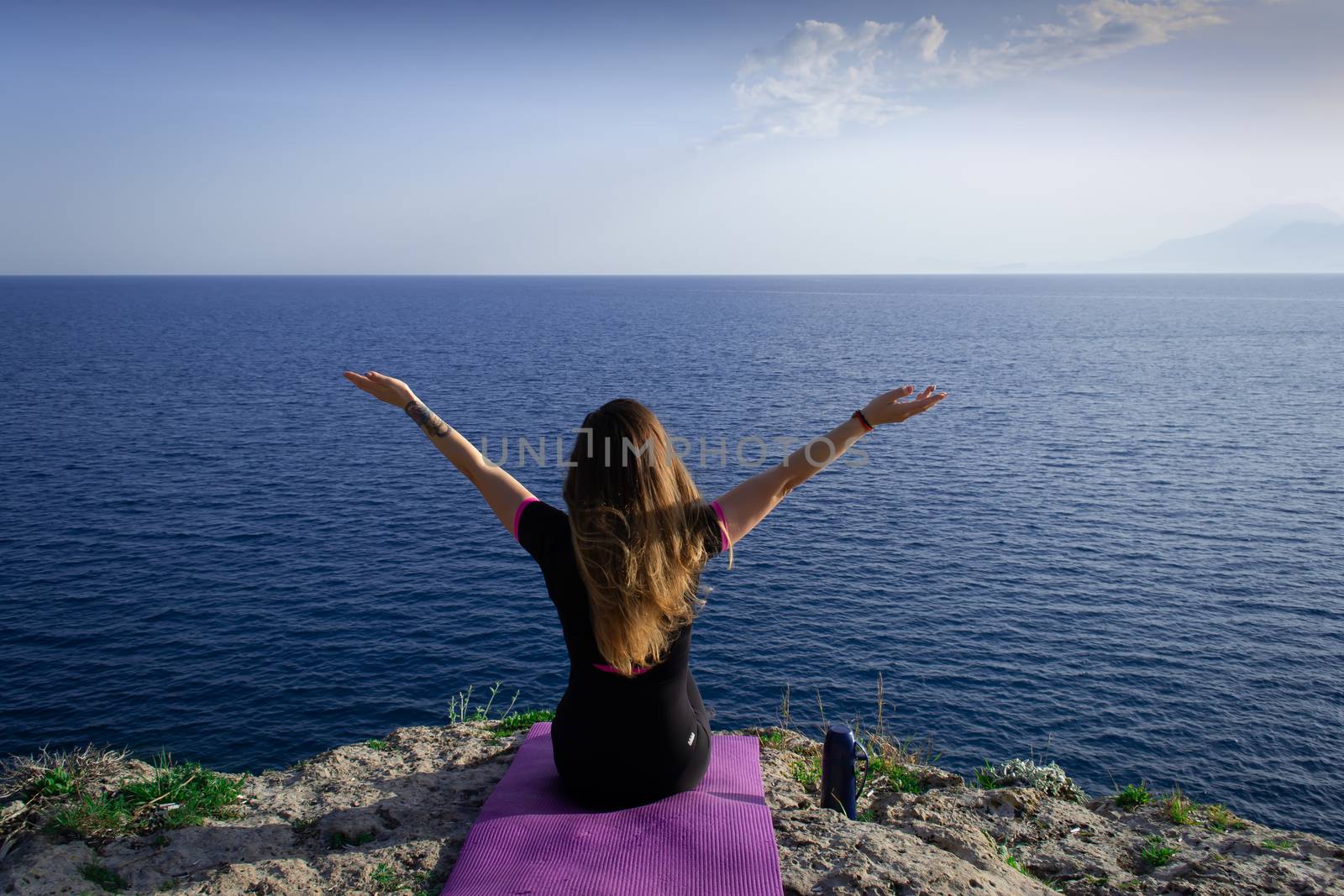 Beautiful young happy lonely girl sitting on rock and watching blue Mediterranean sea and relaxing