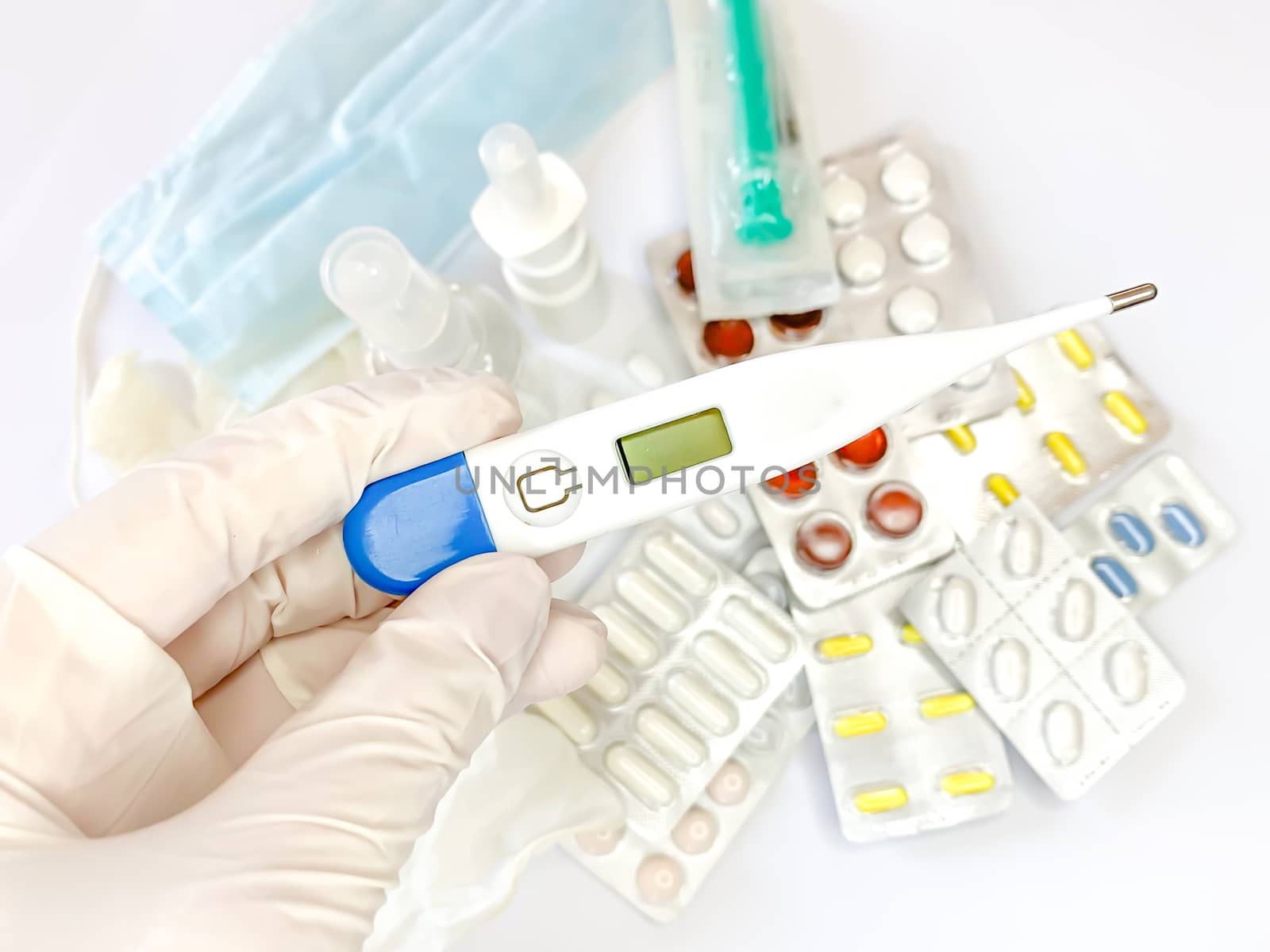 Digital thermometer for measuring body temperature in hand in glove of a woman on white background with many pills and medicines. Horizontal image