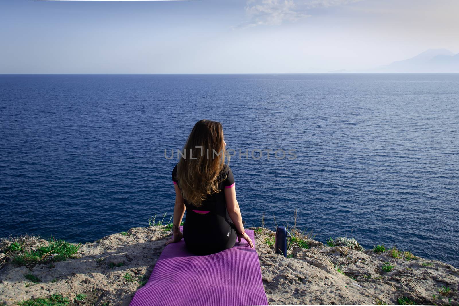 Beautiful young happy lonely girl sitting on rock and watching blue Mediterranean sea and relaxing