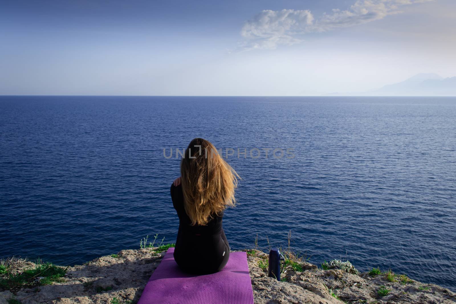 Beautiful young happy lonely girl sitting on rock and watching blue Mediterranean sea and relaxing