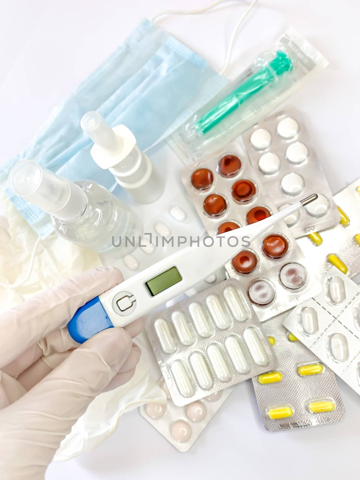 Digital thermometer for measuring body temperature in hand in glove of a woman on white background with many pills and medicines. Vertical image