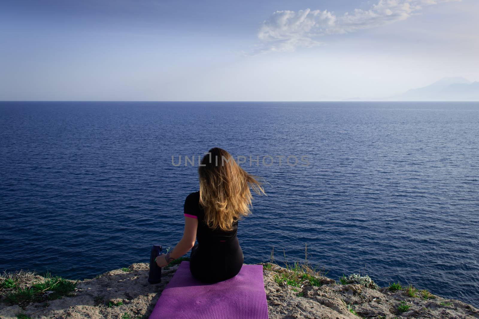 Beautiful young happy lonely girl sitting on rock and watching blue Mediterranean sea and relaxing