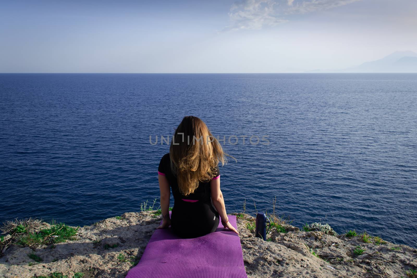 Beautiful young happy lonely girl sitting on rock and watching blue Mediterranean sea and relaxing