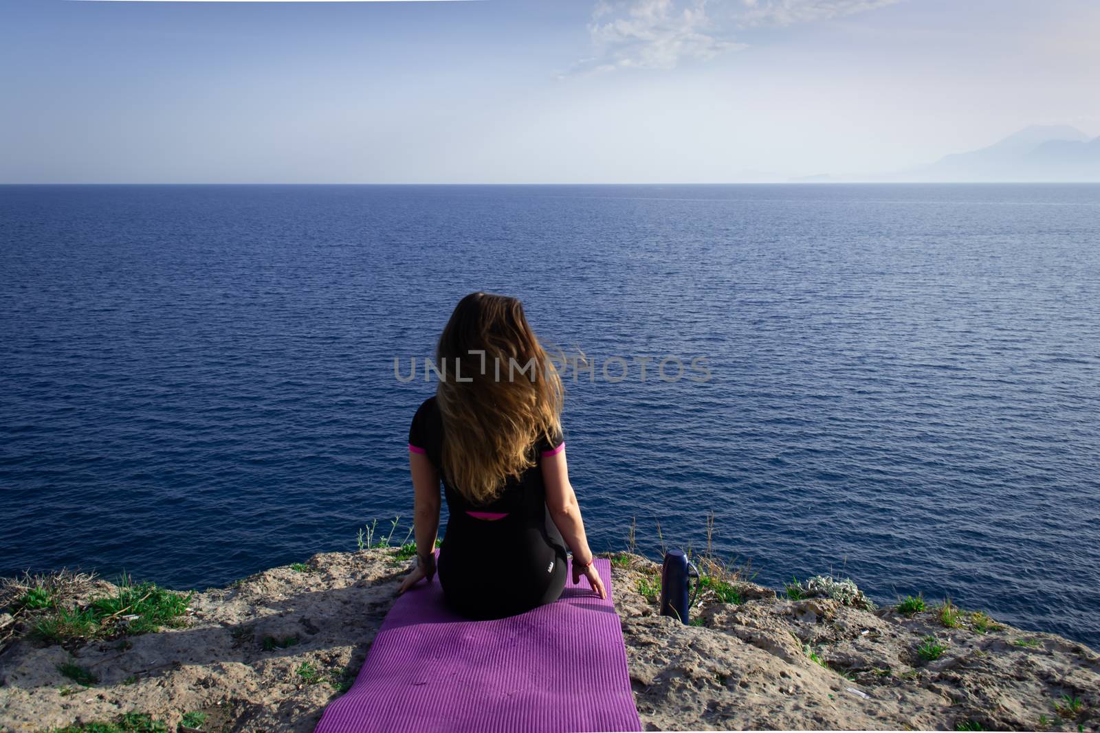 Beautiful young happy lonely girl sitting on rock and watching blue Mediterranean sea and relaxing