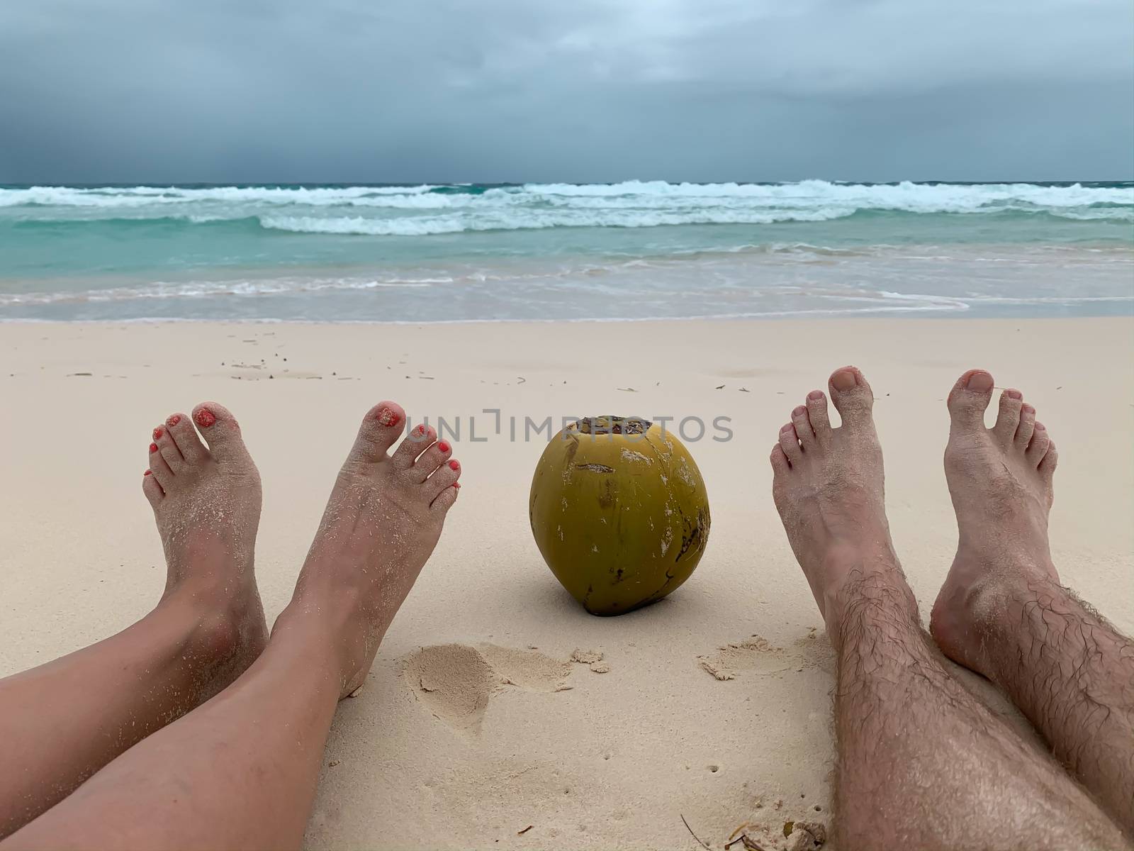 Two pairs of female and male legs with a coconut between them sitting on a white sand Caribbean beach on the ocean background on a cloudy day. Honeymoon image concept.