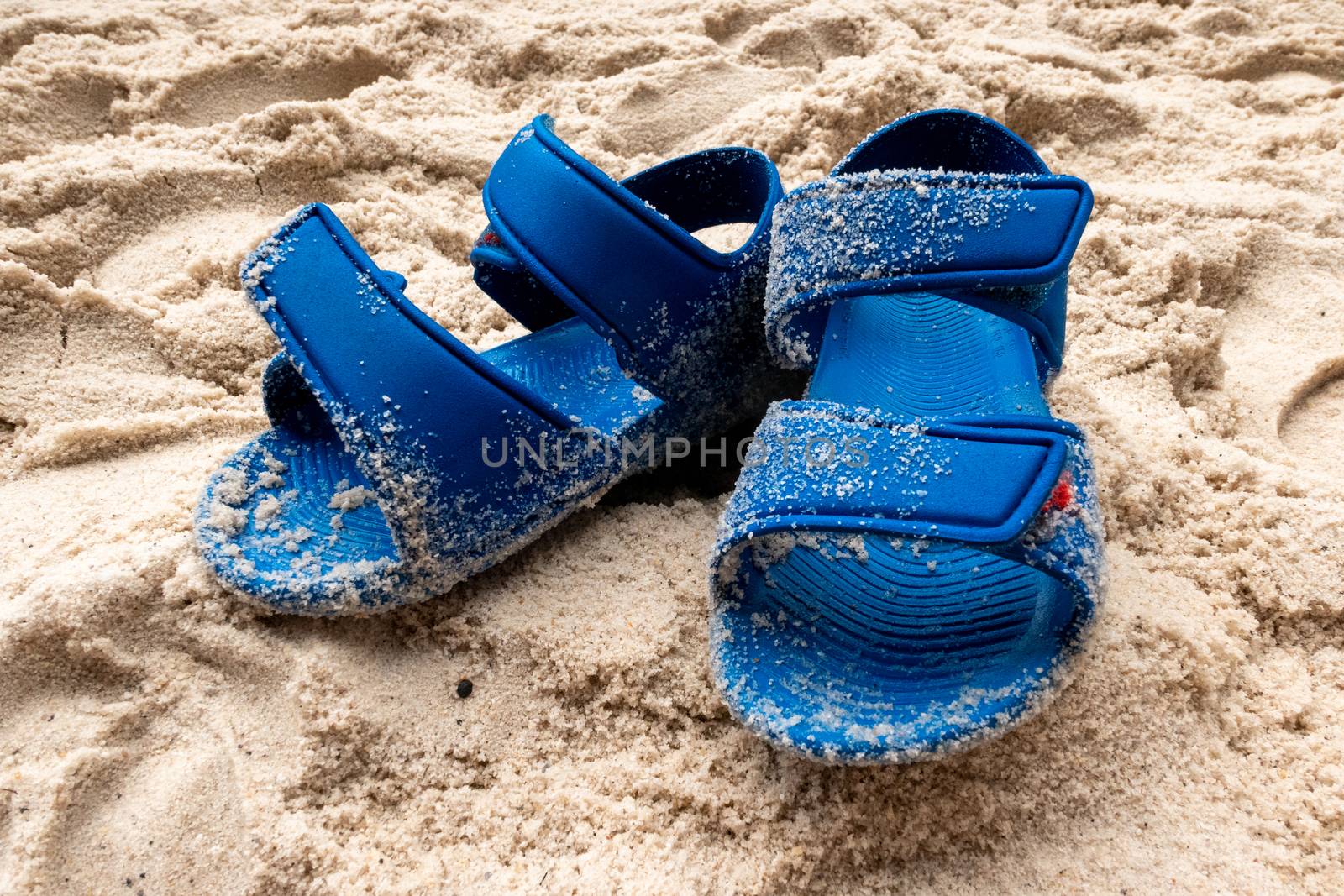 A pair of blue children's sandals standing in the sand on a beach and dirty with sand. The sandals have velcro fastenings.