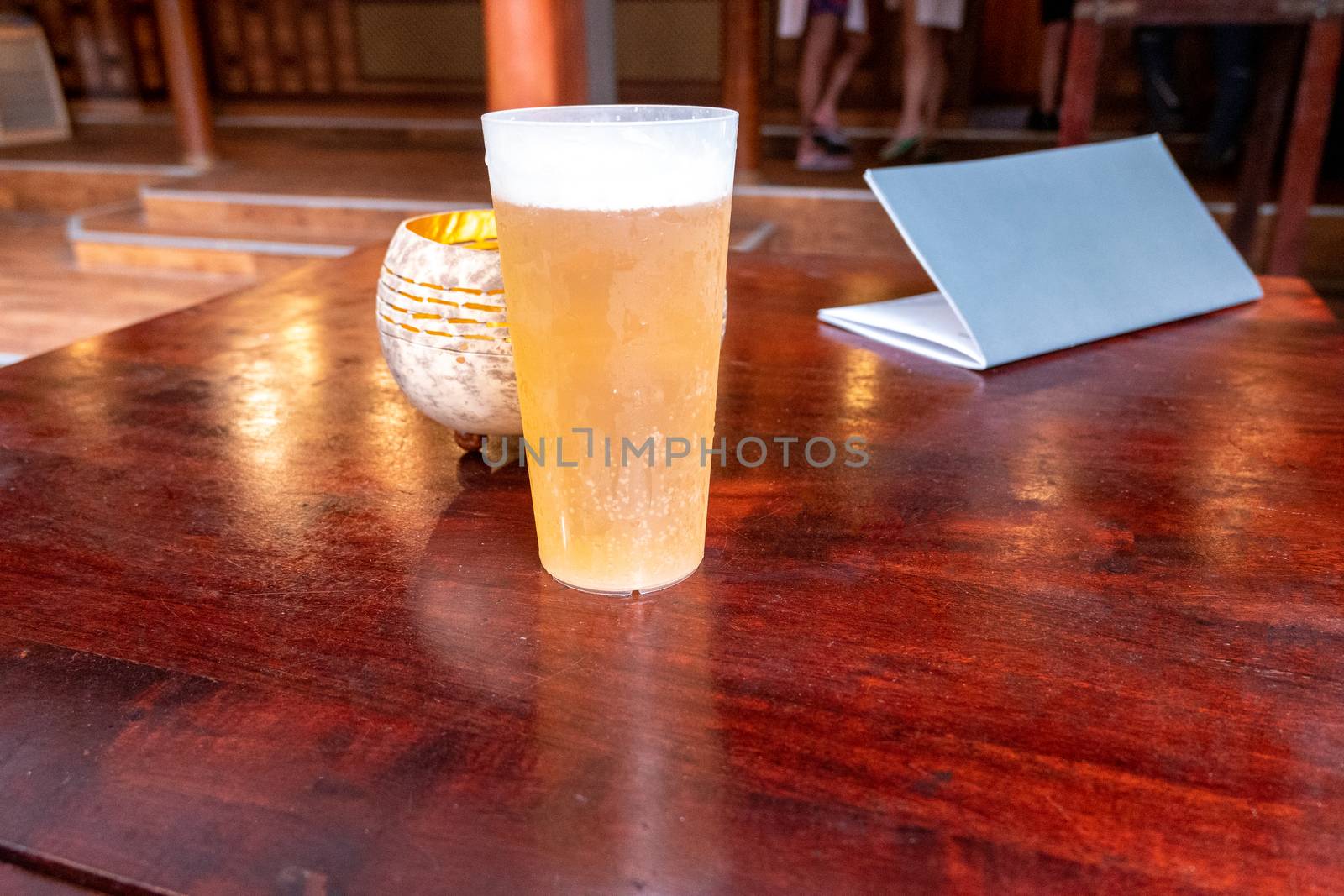 A shiny wooden table in a bar or restaurant with a drink, similar to beer, in a plastic cup. Also a drink or menu card, a gold-coloured bowl for a candle. In the background people at the bar.