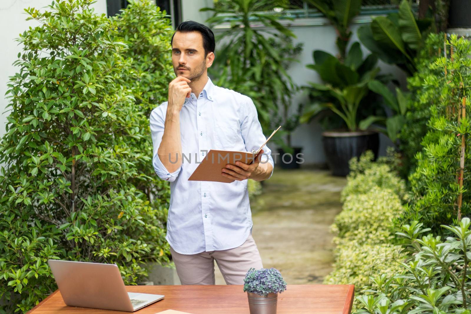 A young man stands reading a book in the garden. There's a notebook on the table to use information and work.