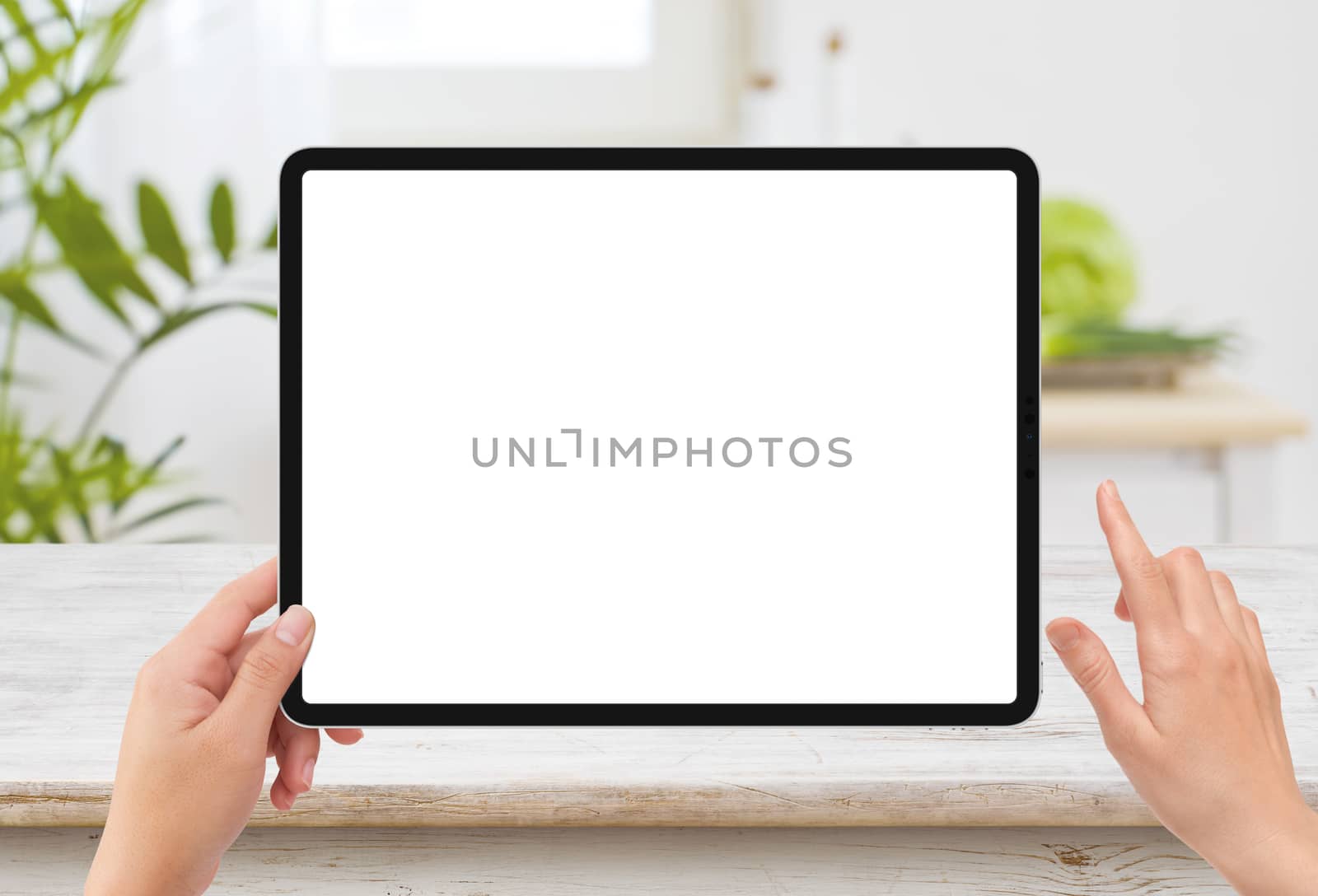 Isolated human left hand holding black tablet media device with white empty screen mockup and wooden table in kitchen