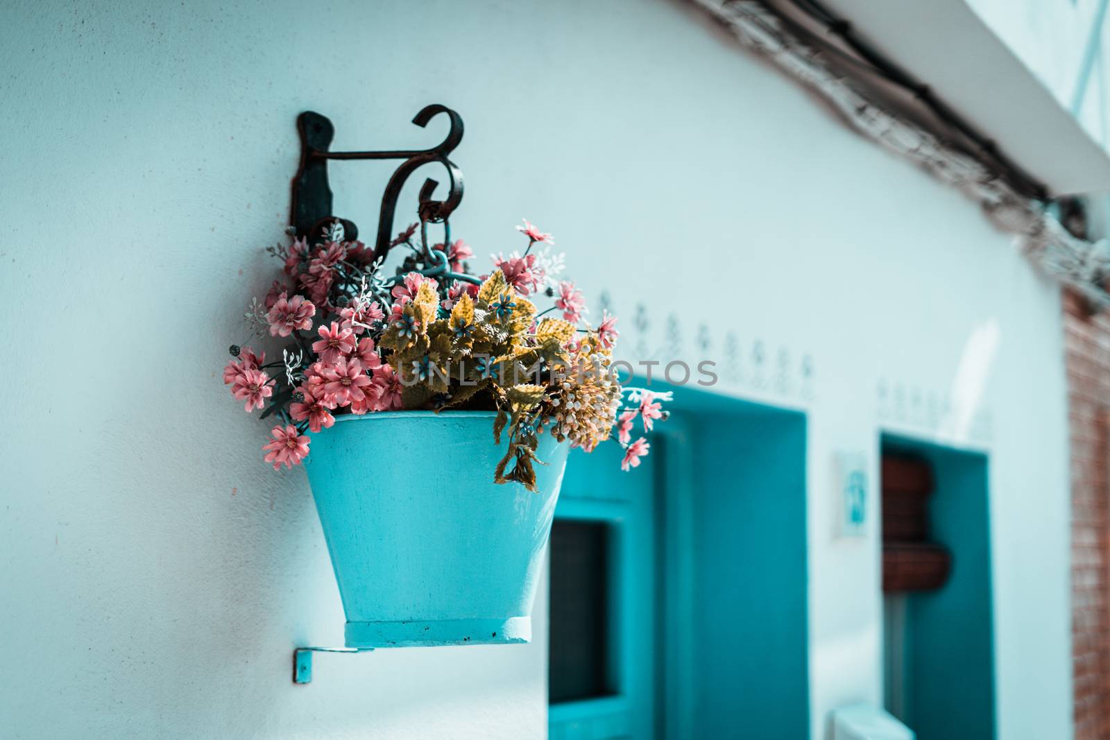 Pink flowers in a blue bucket hung on a white wall in the traditional fishing village of Ametlla de mar. Greece or Ibiza bakground. Teal and orange style