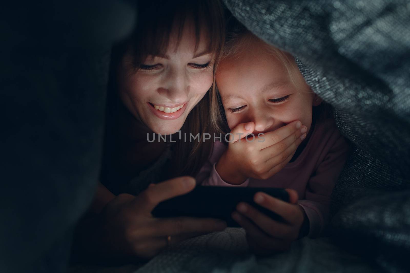 Mother with a little daughter watching content on  smartphone in the dark under the blanket covers.