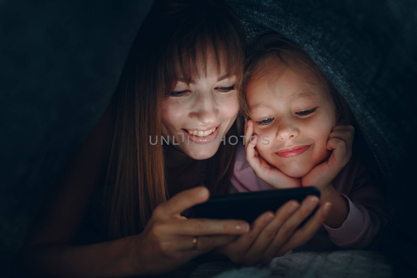 Mother with a little daughter watching content on  smartphone in the dark under the blanket covers.