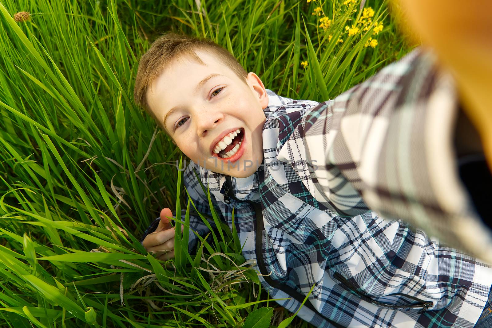 Happy smiling boy making self portrait on smartphone in meadow. young boy making selfie on smartphone laying in green grass with yellow flowers