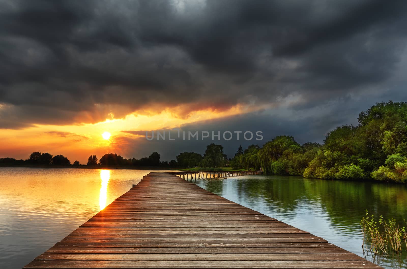 Wooden path bridge over lake at stormy dramatic sunset by necro79