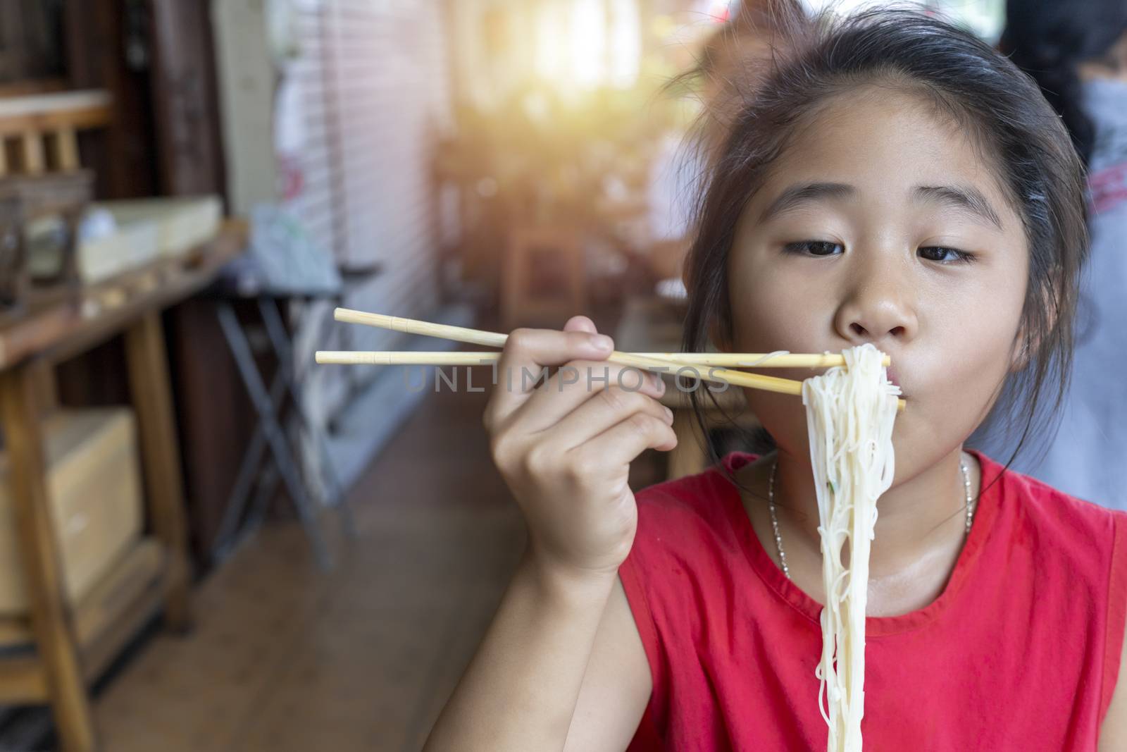 Asian child girl with red dress eating small noodles soup with chopsticks at restaurant