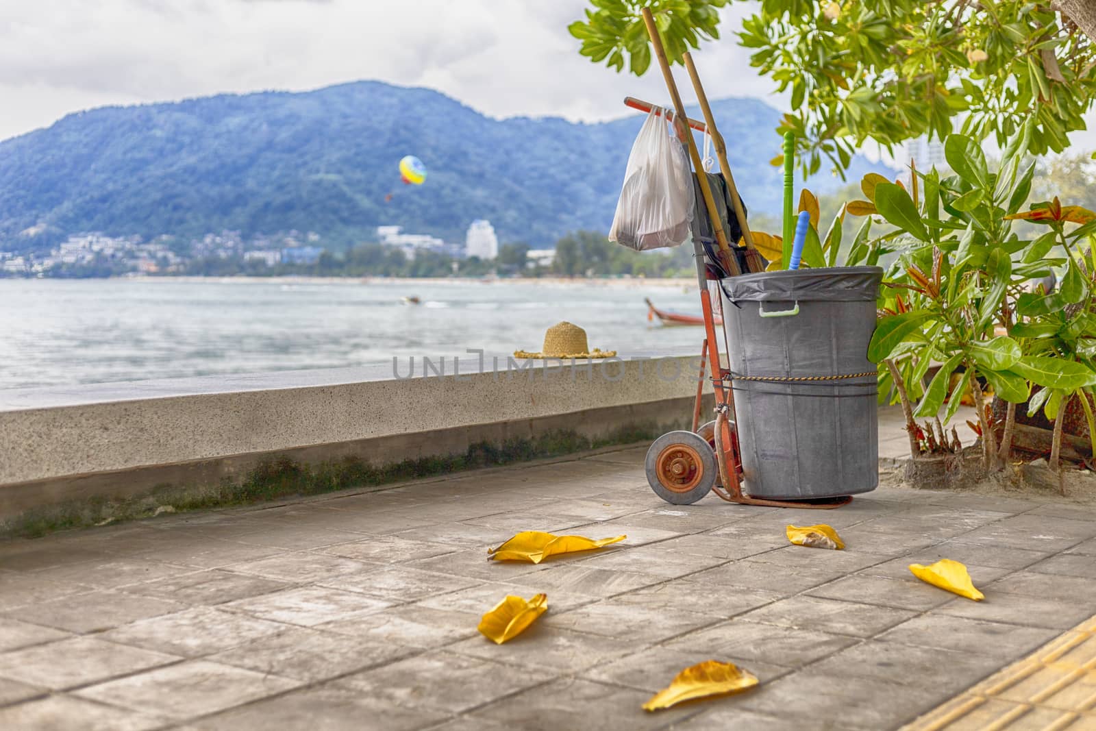 Black bin on Cart with dust pan and plastic garbage bag stay on sidewalk under the tree near the beach.