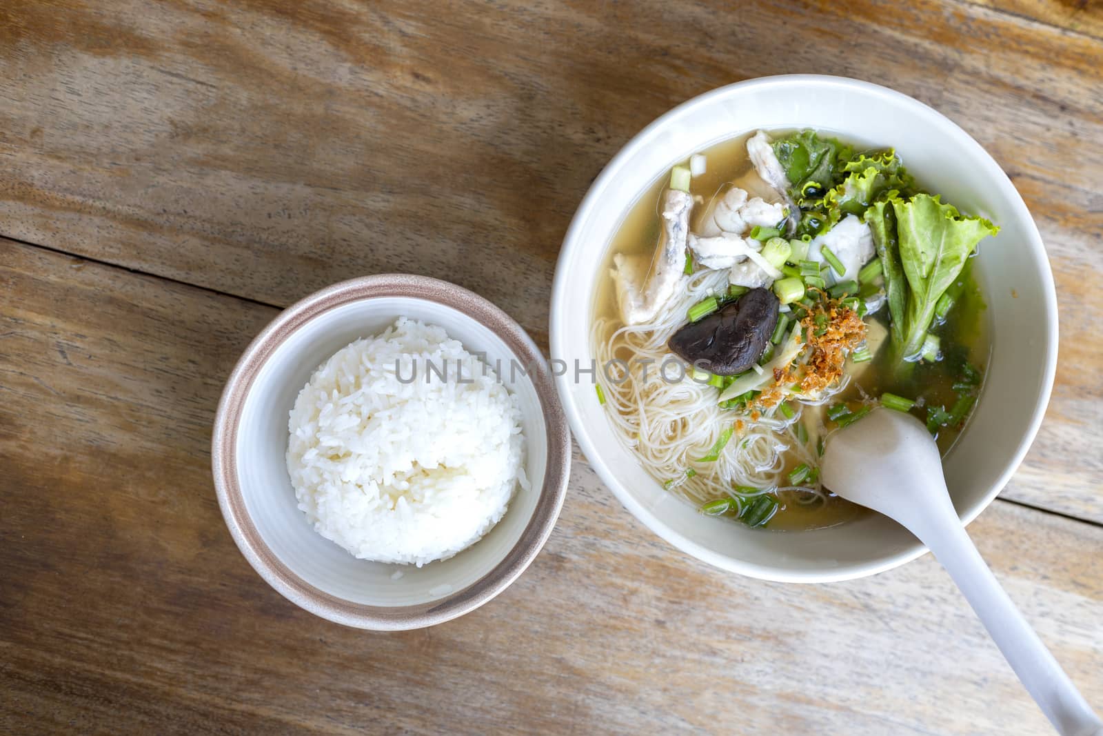 Soup with thin noodle, pock, pock stick, mushroom, vegetable in white bowl near have rice in small bowl on wood table. with copy space for text or design