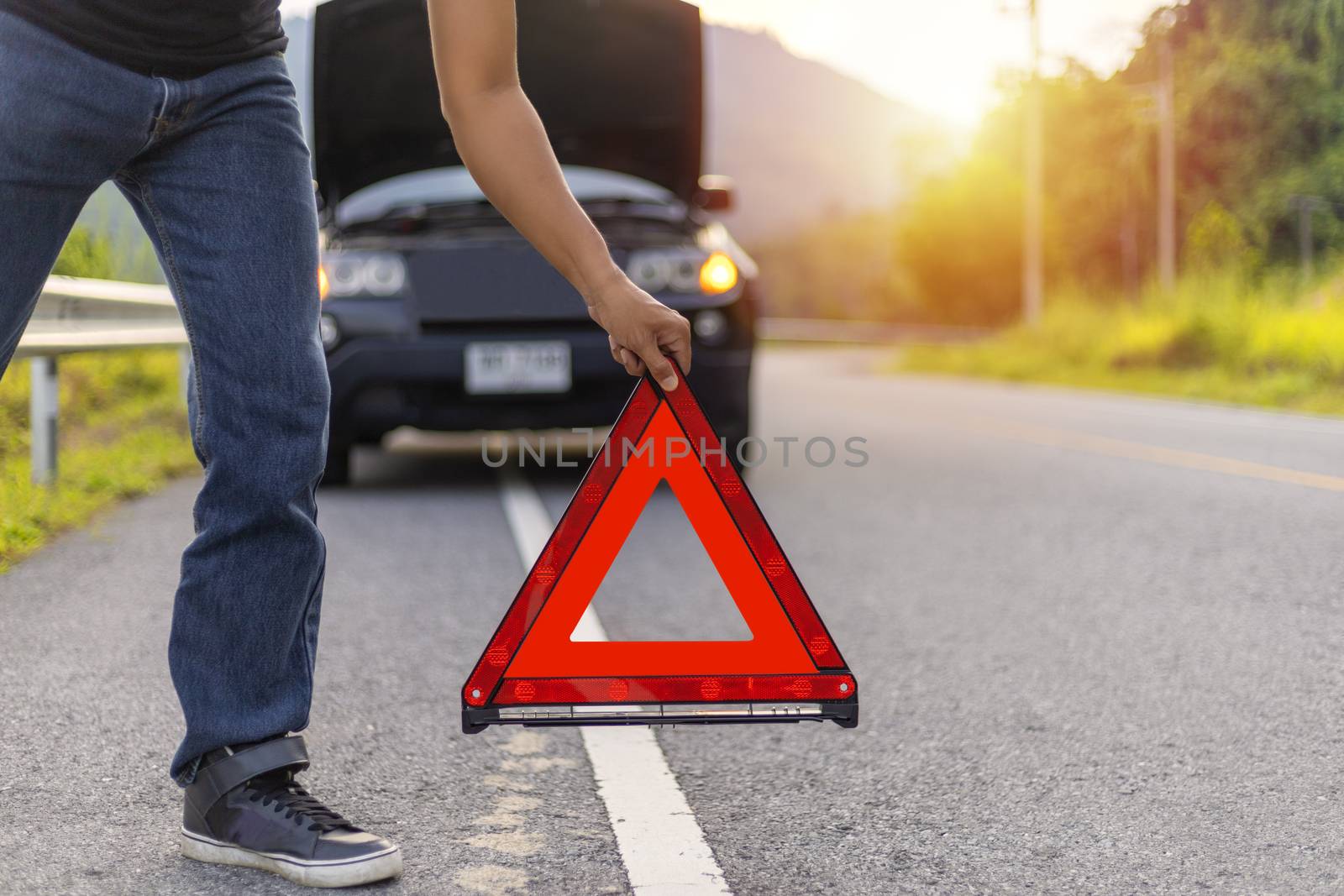 Man hold and put down the red triangle for warning have black color car parking  on road and oper car hood.